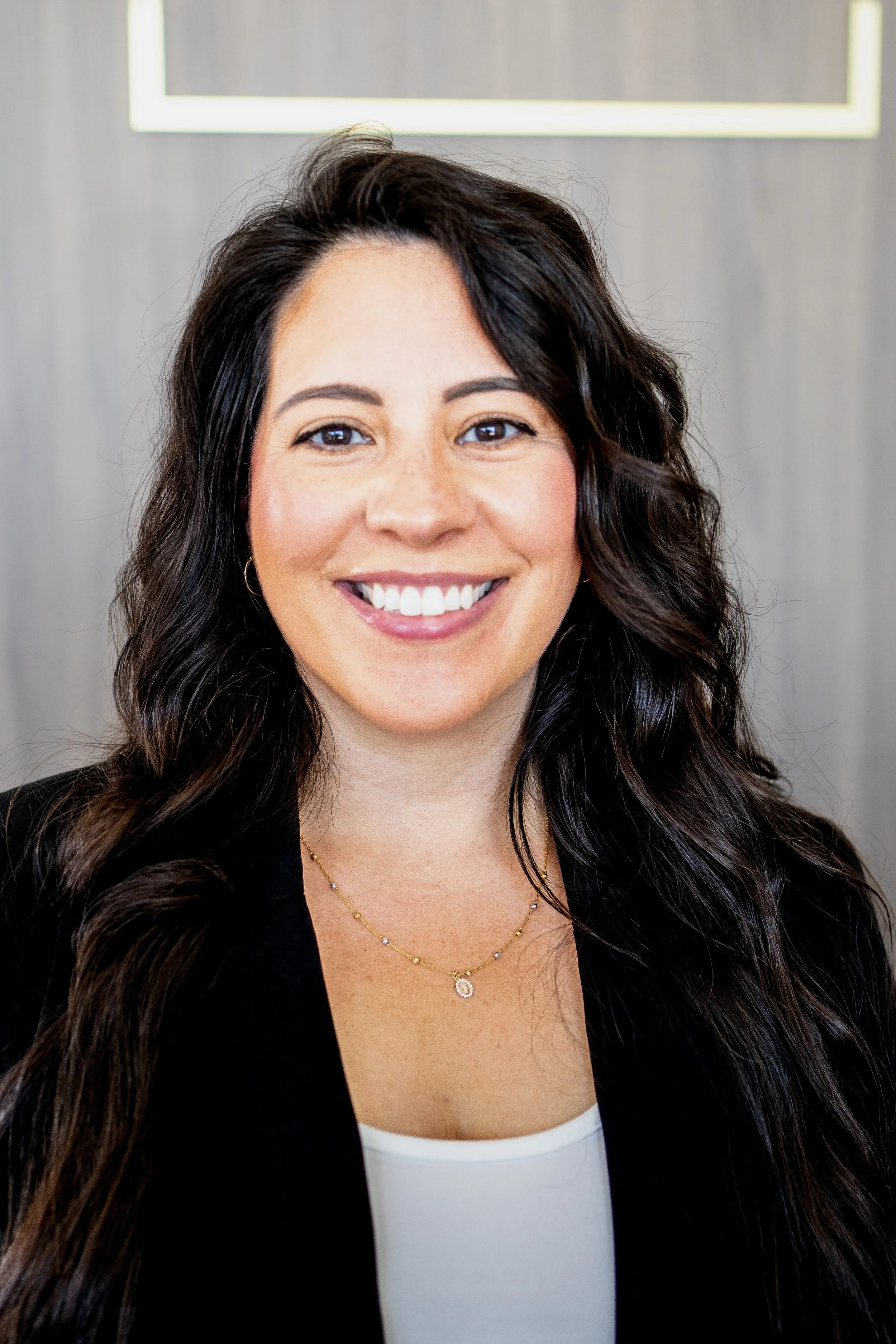 Close-up portrait of a smiling woman with dark, wavy hair, wearing a black blazer, white top, gold necklace, and earrings, against a gray background.