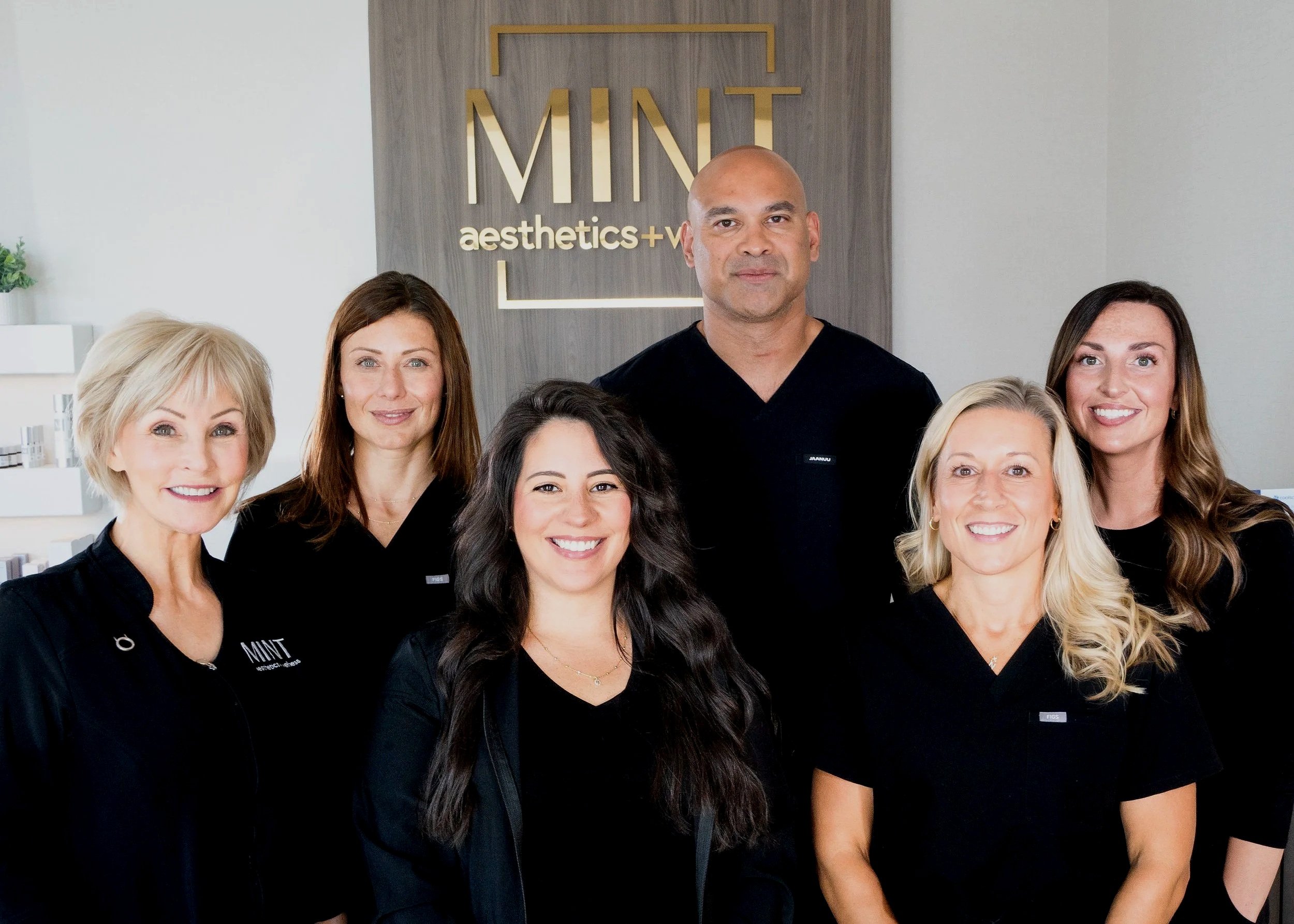 Group of diverse women and a man in black professional attire standing in front of a wall with the logo