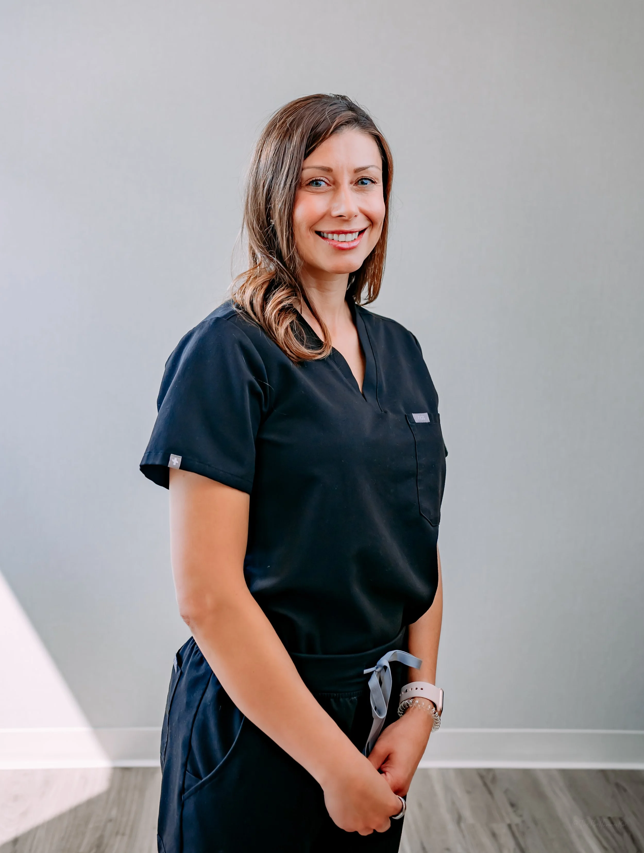 Smiling woman in black scrubs standing indoors against a gray wall.