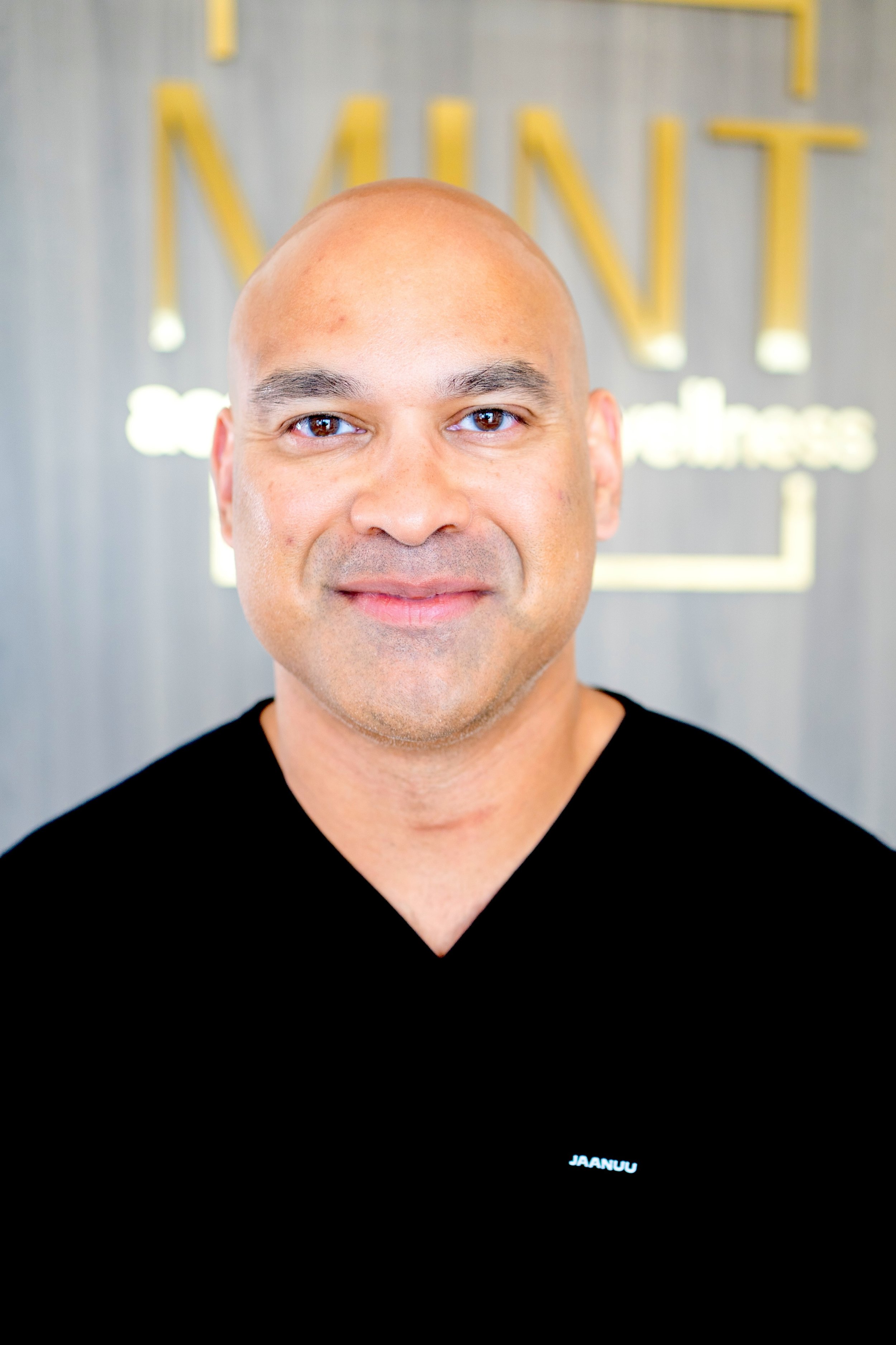 A man with a bald head, dark eyebrows, and a light brown complexion smiling at the camera, wearing a navy blue medical scrubs and a silver wristwatch, standing against a light gray wall.