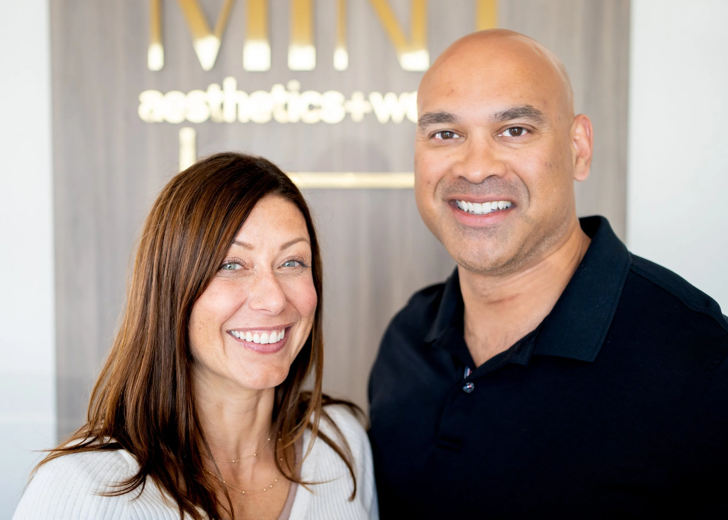 A close-up selfie of a smiling middle-aged woman with long brown hair and a man with a shaved head, both standing in front of a sign that reads 'aesthetics + wellness'.