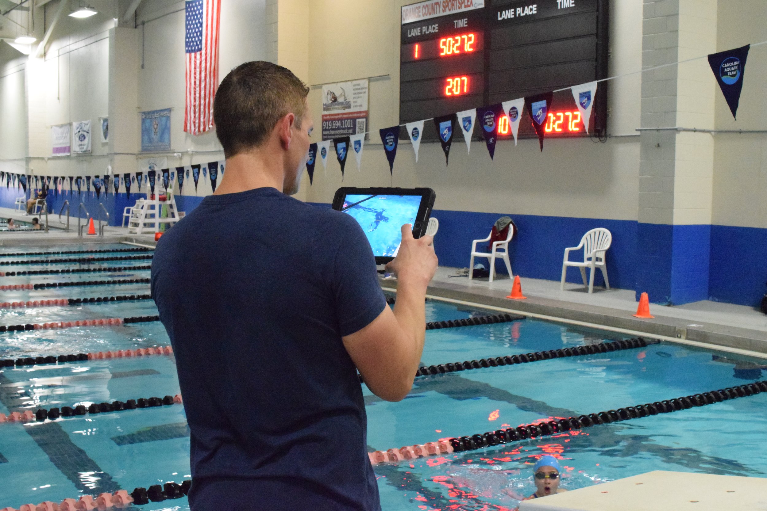 A swimmer in a pool is being observed or coached by a man using a tablet device. The background features a digital timer board showing elapsed time and lap count, and there are swimming flags and other swimmers around the pool.