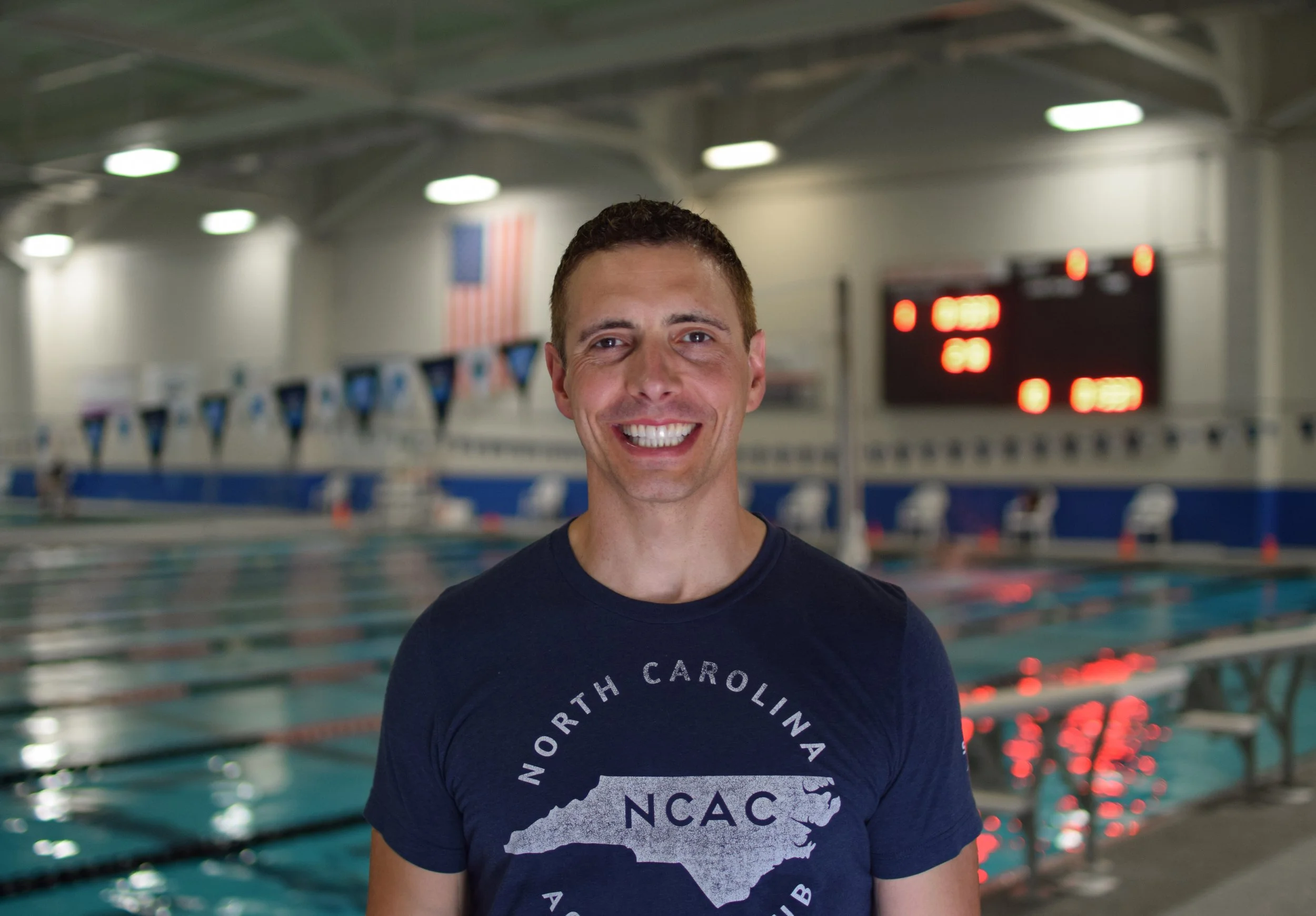 Smiling man with short, dark hair standing in front of a swimming pool at an indoor aquatic center. He is wearing a navy blue T-shirt with the words North Carolina in a circular pattern and a graphic of the state. In the background, there are flags, swim lane markers, and a digital scoreboard.