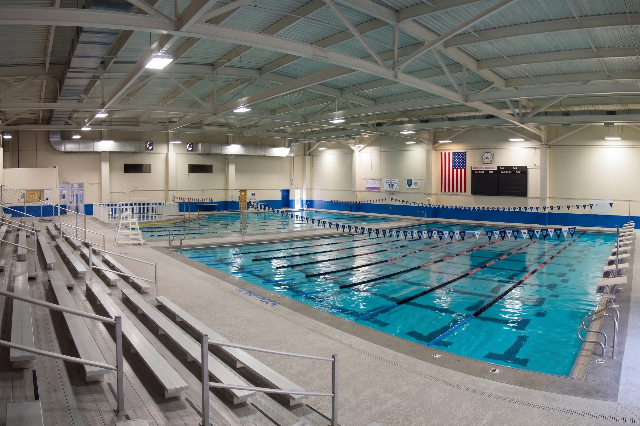 Indoor swimming pool facility with multiple lanes, bleachers, American flag, and scoreboard, under a high ceiling with metal beams and bright lighting.
