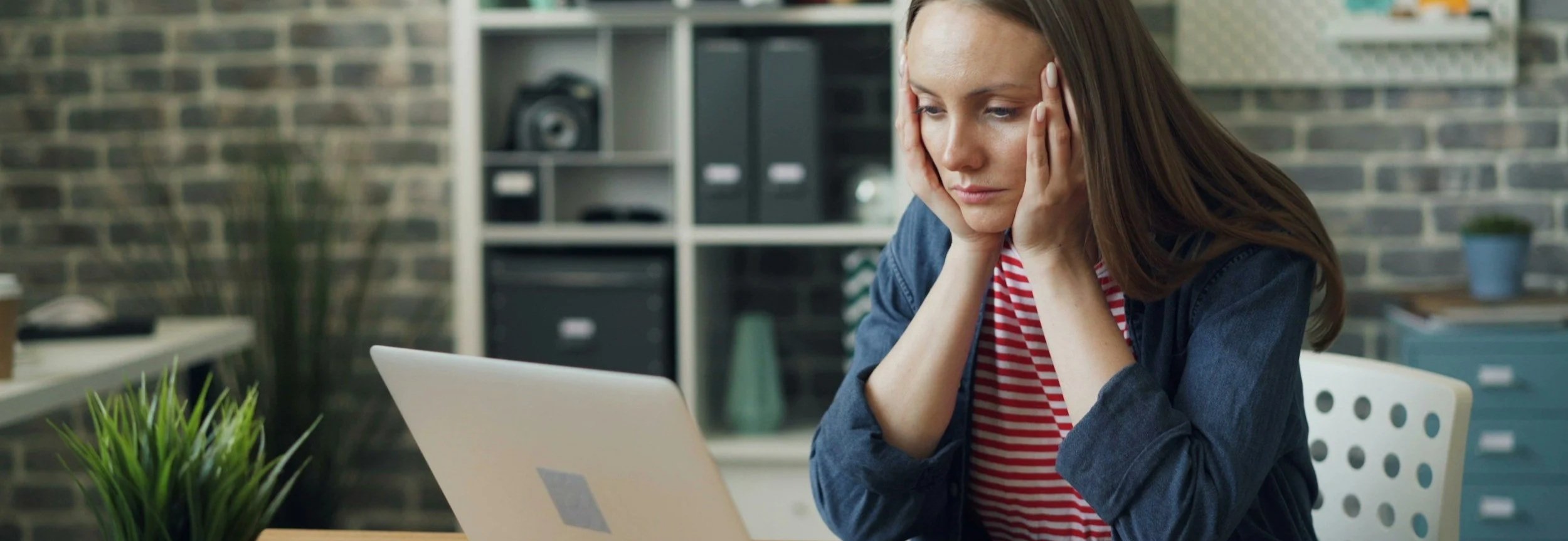 Woman sitting at a desk, holding her face looking stressed while staring at a laptop.
