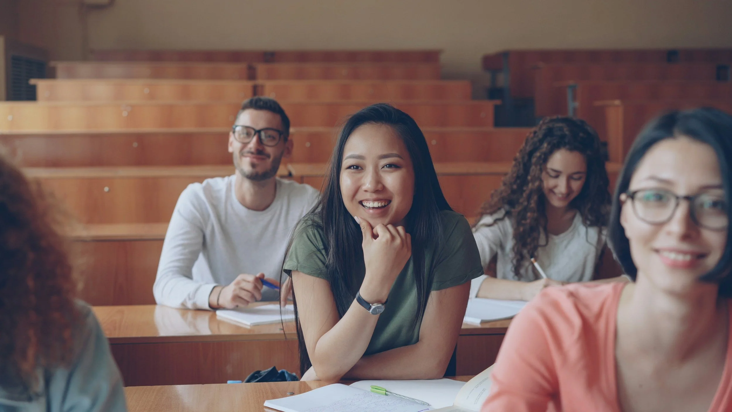 A group of diverse young students sitting in a classroom with wooden tiered seating, smiling and engaging in a discussion, with notebooks and pens on the table.