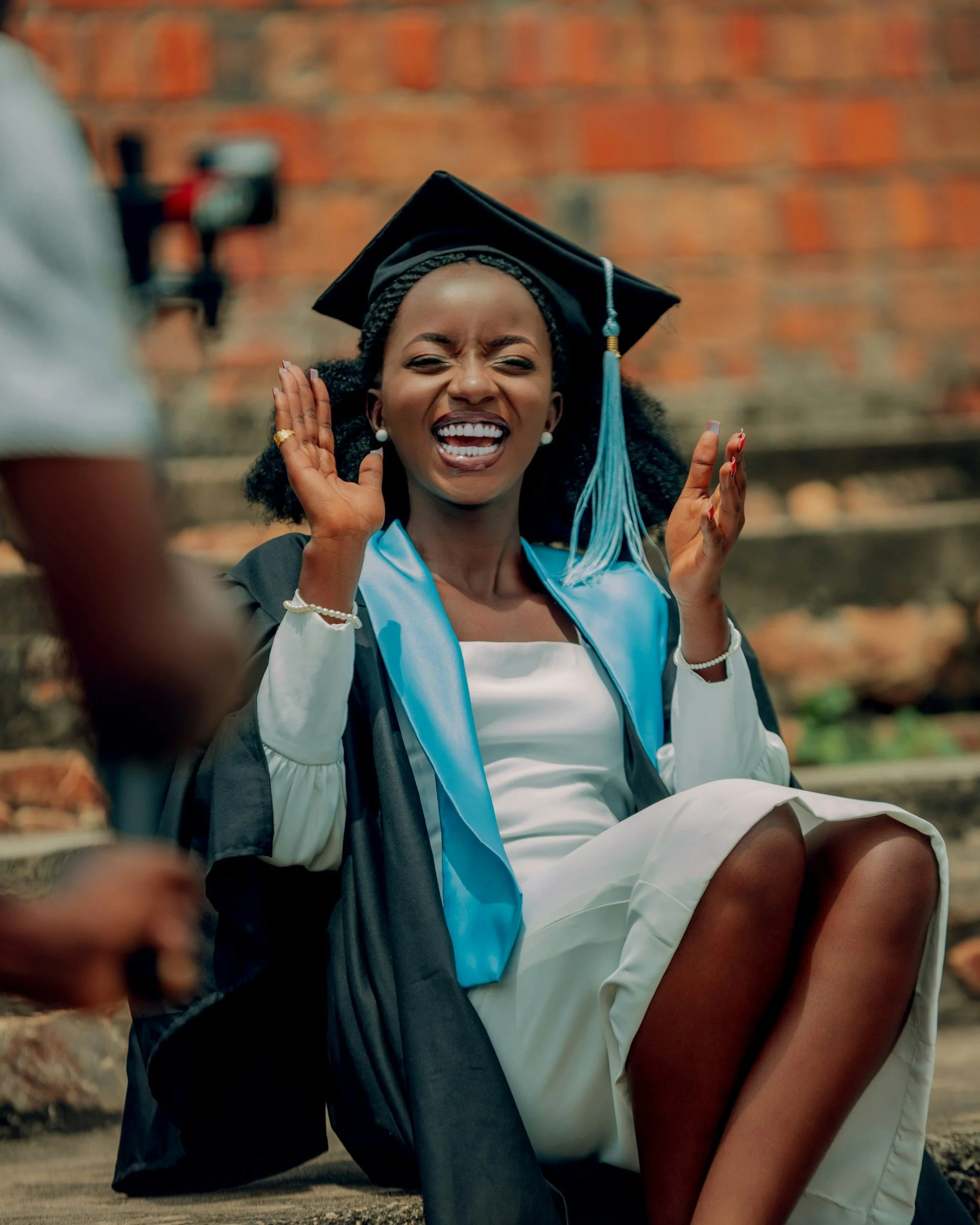 Black woman sitting down in a formal outfit with a grad cap and gown on, smiling and raising her hands as if she's about to clap
