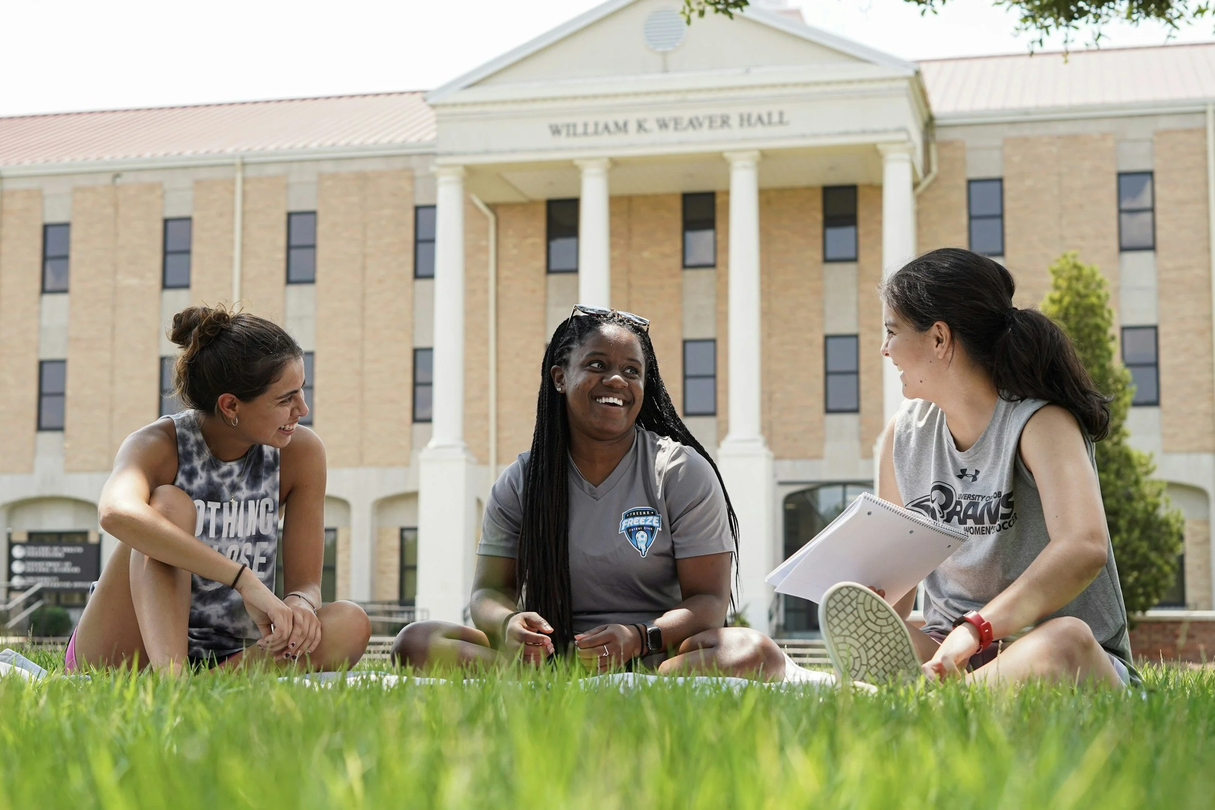 Two female students sitting outside of a university building on the grass, smiling at eachother while one is holding a notebook