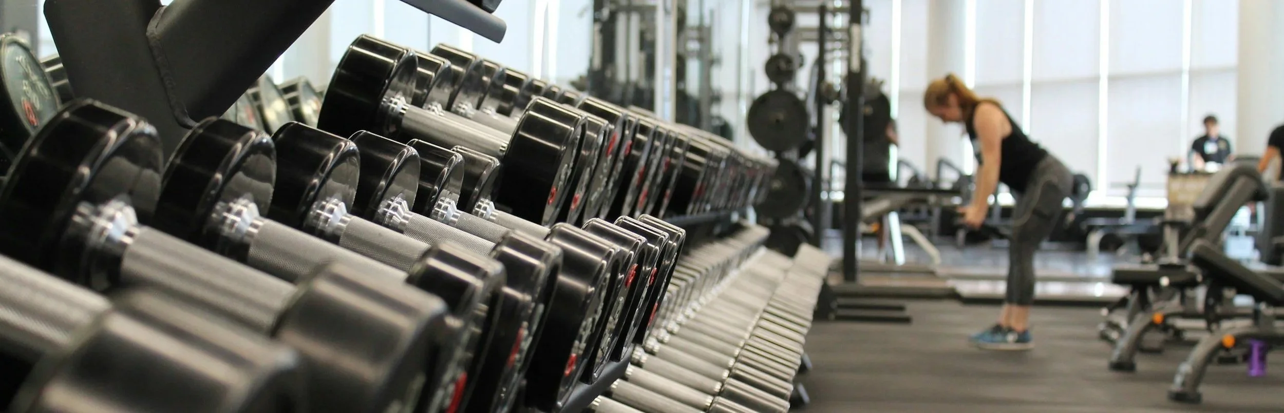 Row of dumbells at a gym with a woman lifting weights in the background