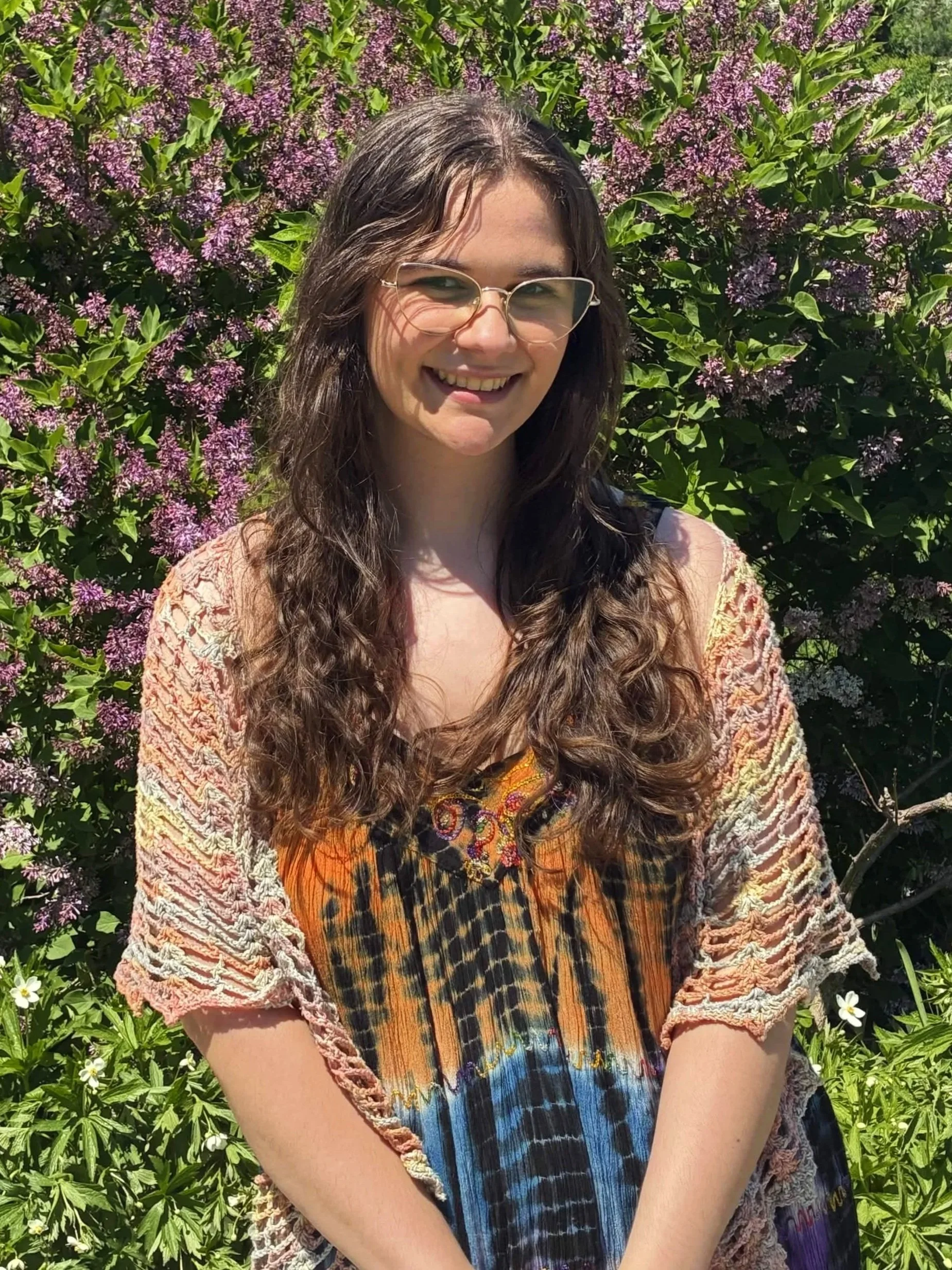 A young woman with long curly hair and glasses smiling outdoors in front of a large bush with purple flowers and green leaves.