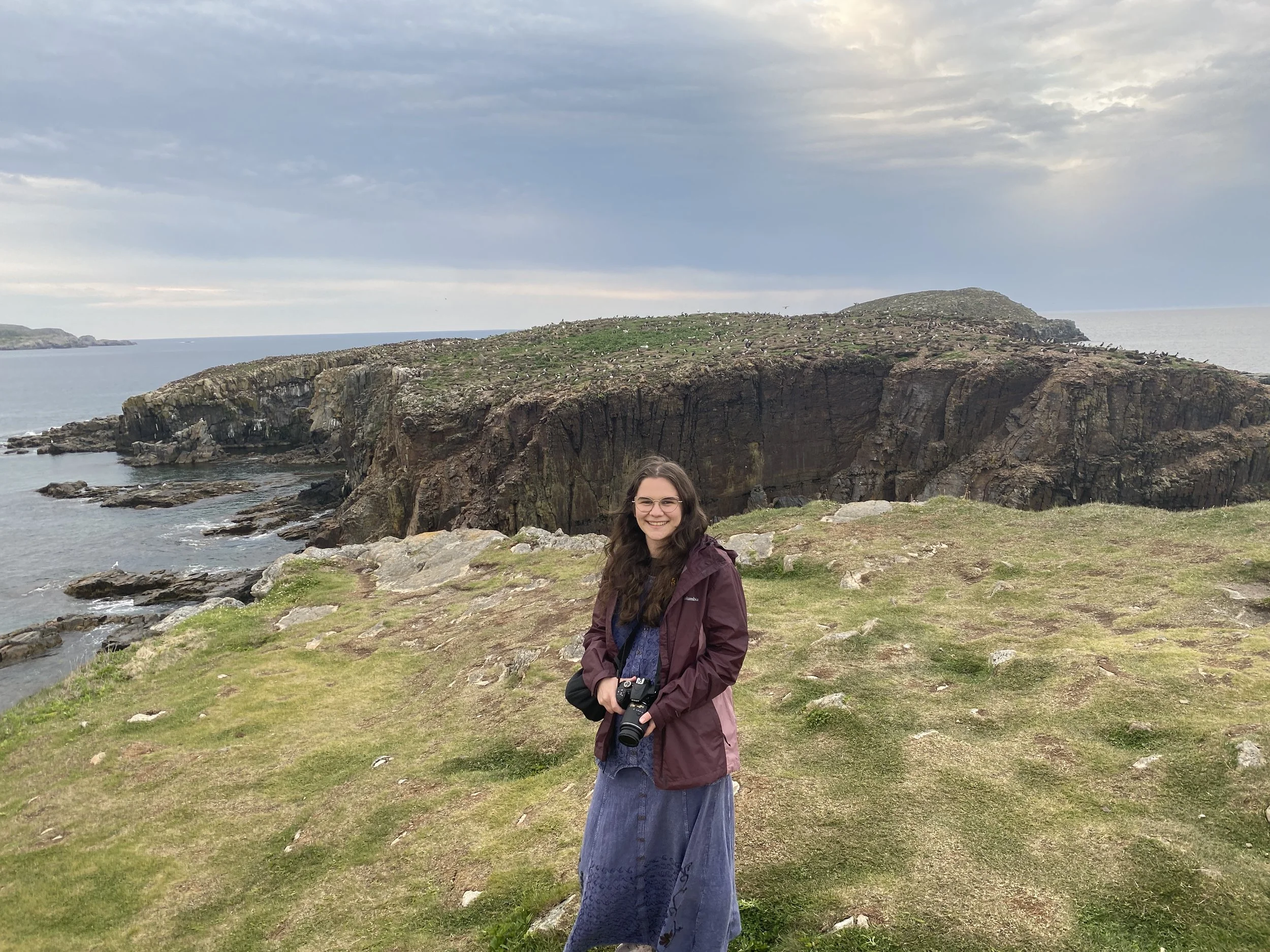 Zoe Lewis, certified ADHD coach in Toronto, a white woman with long wavy brown hair and glasses.  She is holding a camera, and standing on a rocky cliff in Newfoundland.
