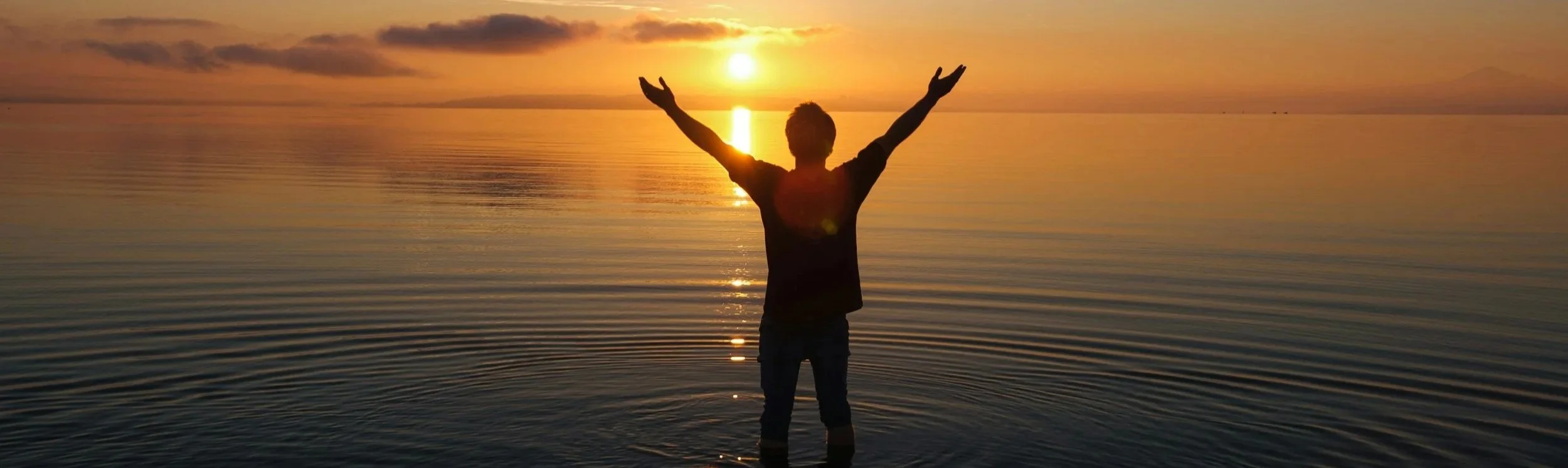 Person standing in water with their arms up at sunset