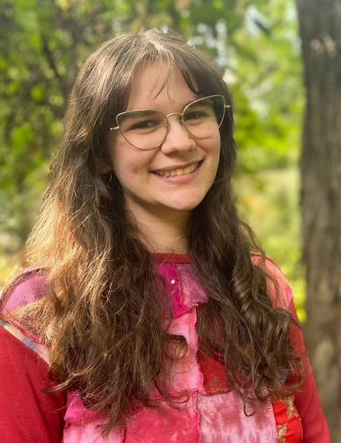 Photograph of Zoe, a white woman with wavy brown hair, cateye gold glasses and a red and pink patchwork top.