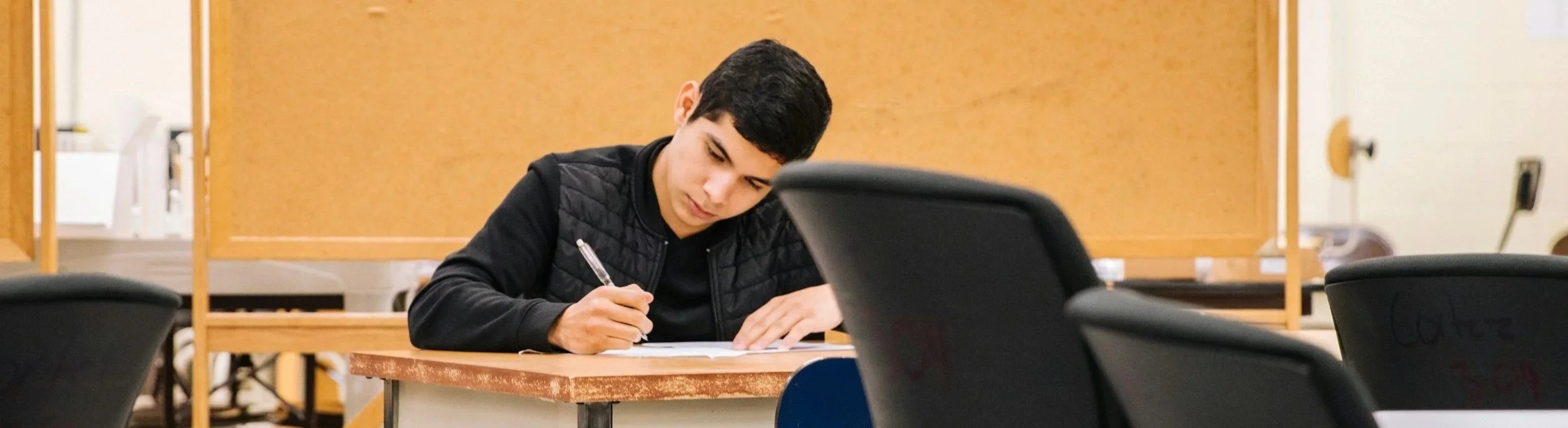 Male student sitting at a desk in a classroom, looking down at a piece of paper and holding a pen.