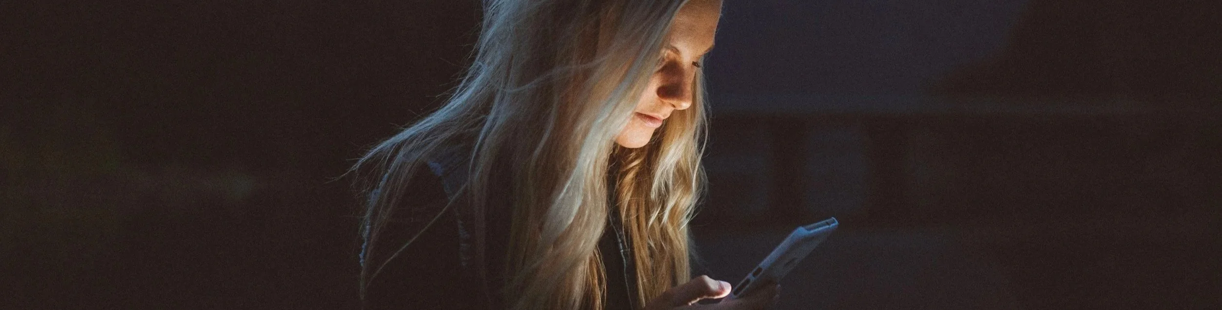 A woman starting at the screen of a smart phone in the dark, her face being lit up by the light.