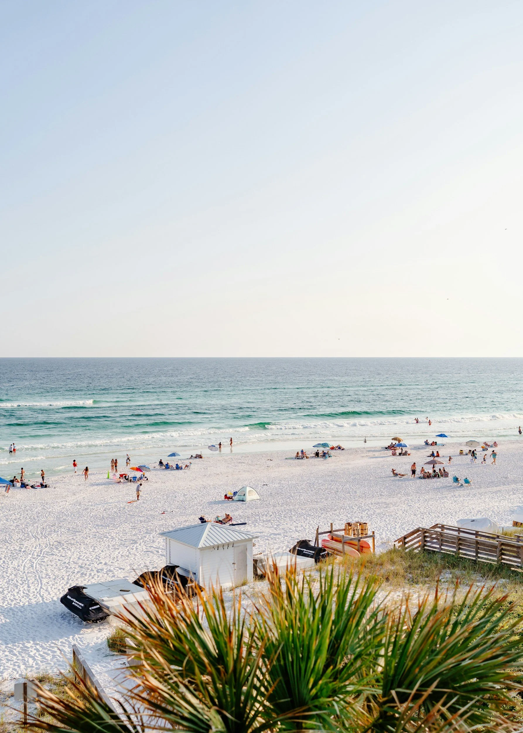 Beach scene with white sand, ocean waves, and people relaxing under umbrellas, with small structures and greenery in the foreground Gulf Coast