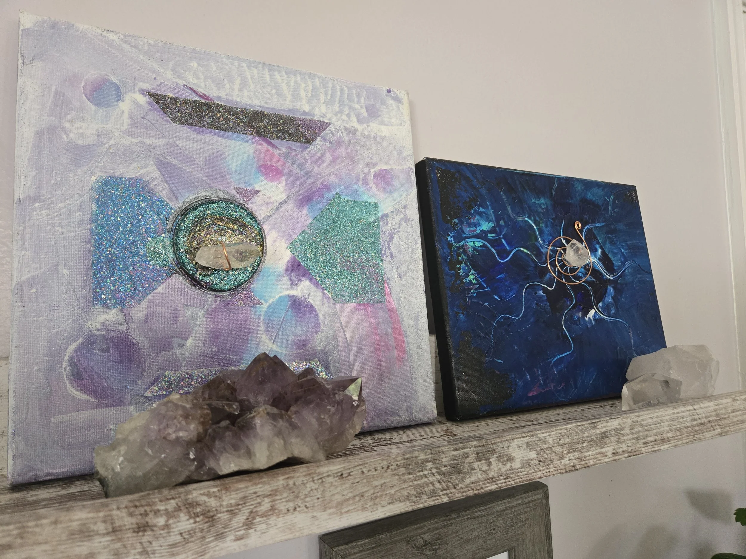 Two abstract paintings with cosmic themes, featuring glitter and swirling patterns, displayed on a wall. The left painting is primarily in purple and pink tones with geometric shapes, and the right painting is predominantly dark blue with swirling lines. There are mineral specimens in front of the paintings, resting on a rustic wooden surface.