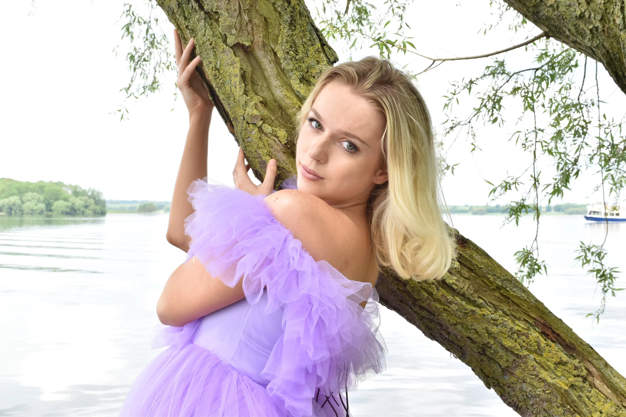 A woman with blonde hair holding onto a tree by a lake, wearing a purple ruffled dress, with water and sailboats in the background.