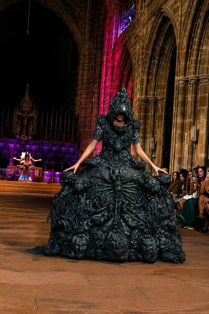 A model wearing an elaborate black dress with intricate textures walks on a runway inside a large, gothic cathedral-style hall with high arched ceilings. Audience members sit along the side, watching and taking pictures.