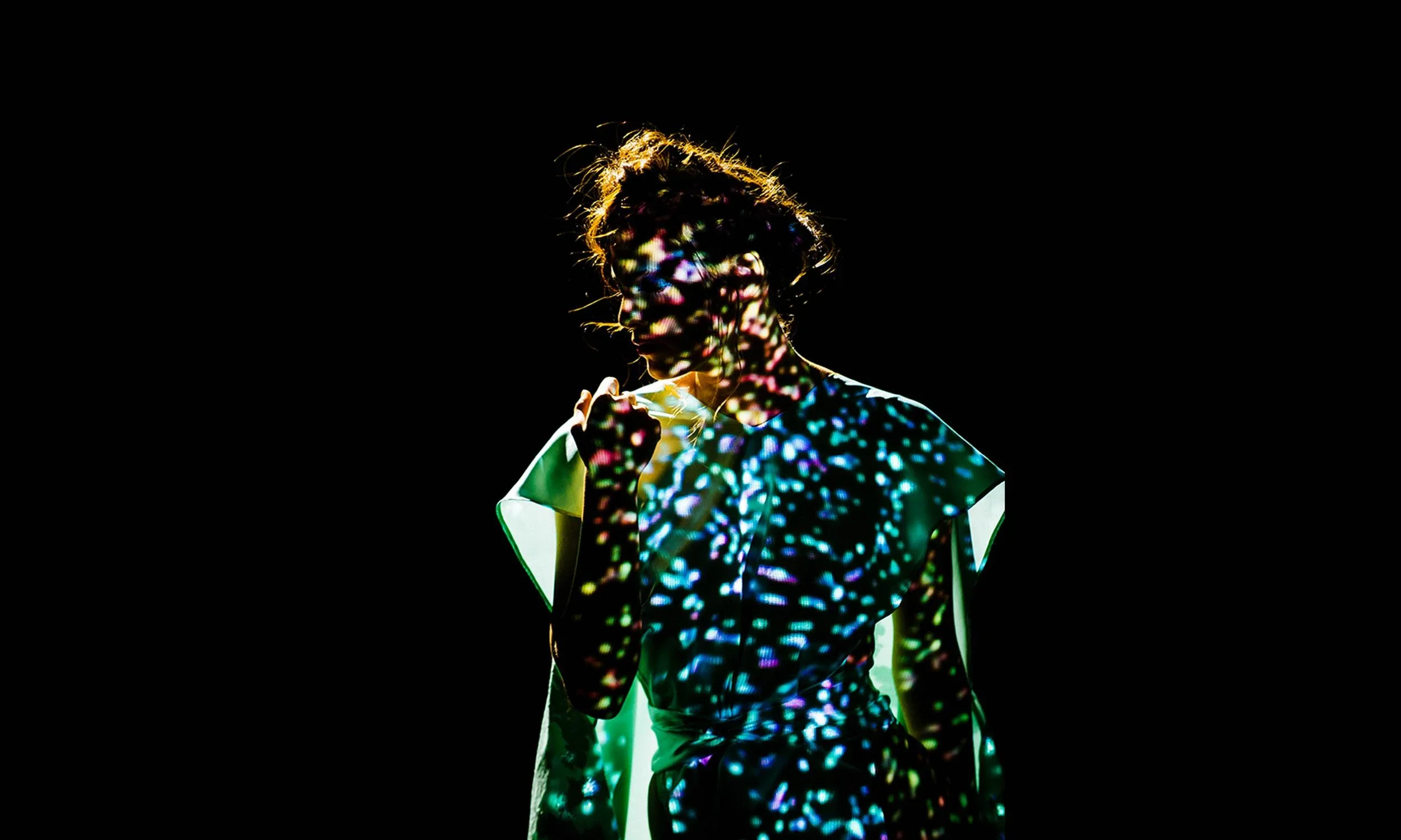 A person with short hair holding their collar, with colorful light projections on their face, hair, and clothing, against a black background.