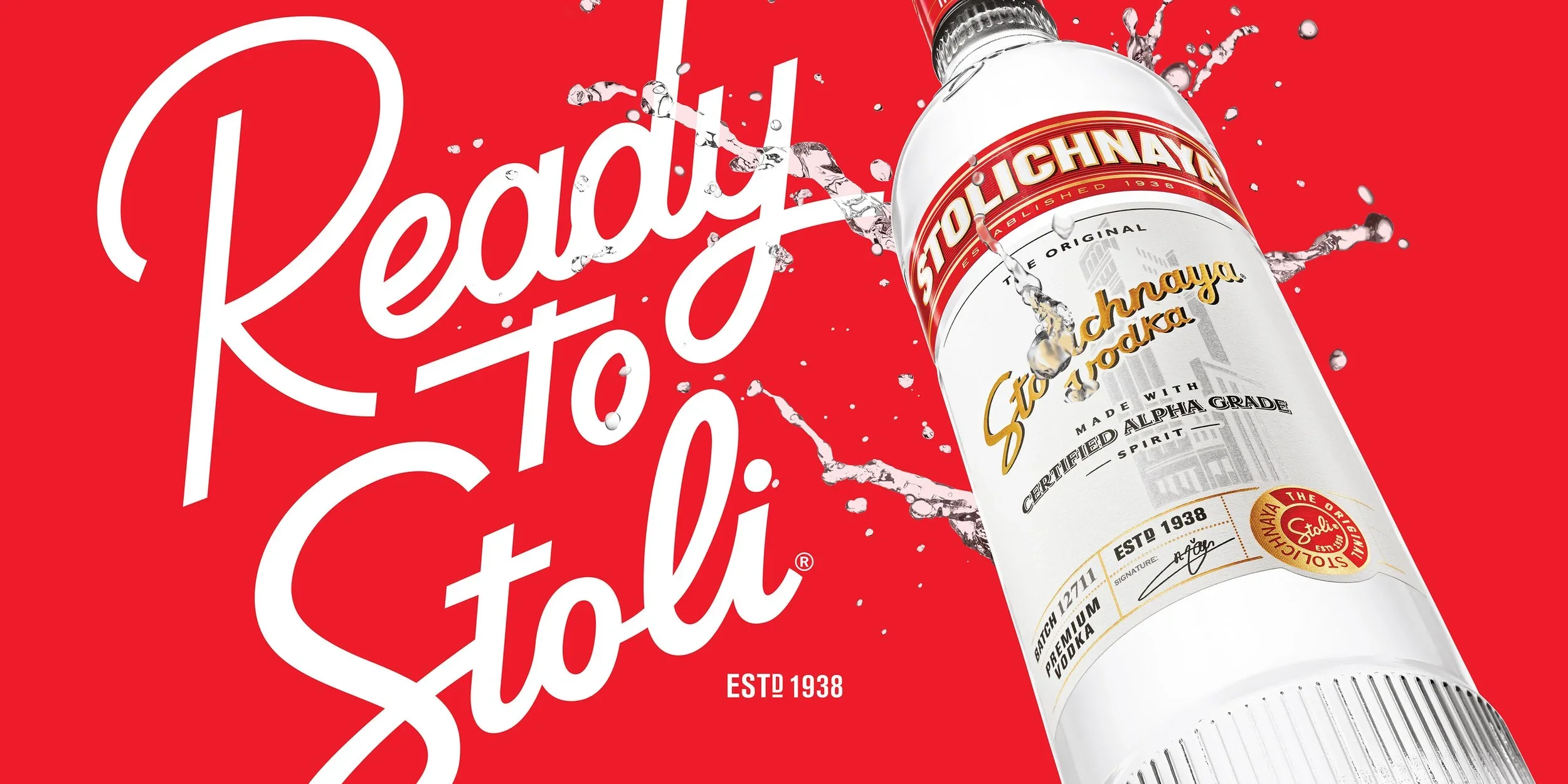 A bottle of Stolichnaya vodka with splashes of water around it on a red background with the Stolichnaya logo and brand information.
