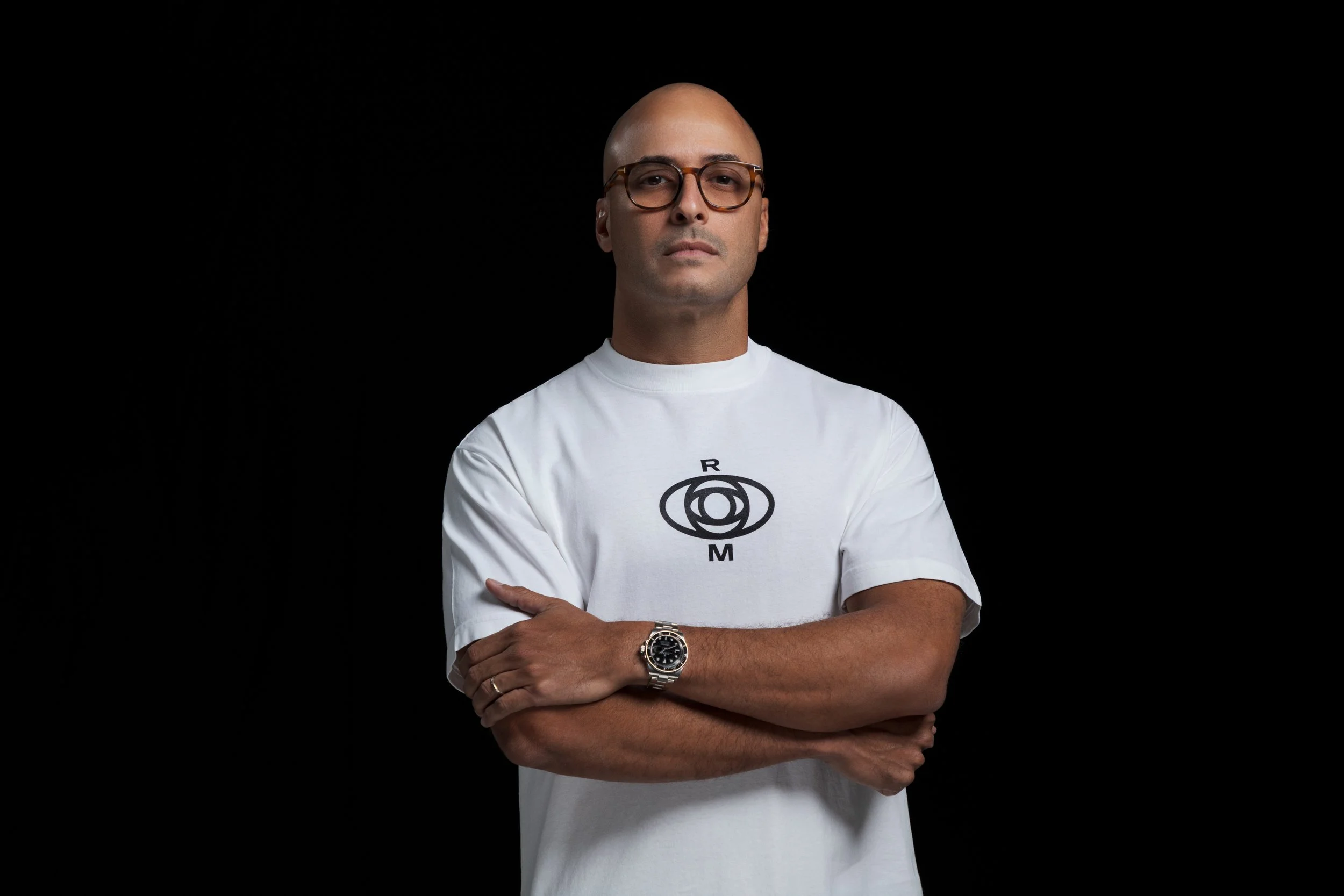 A man with a shaved head and glasses, wearing a white t-shirt with a circular logo, crossing his arms and standing against a black background.