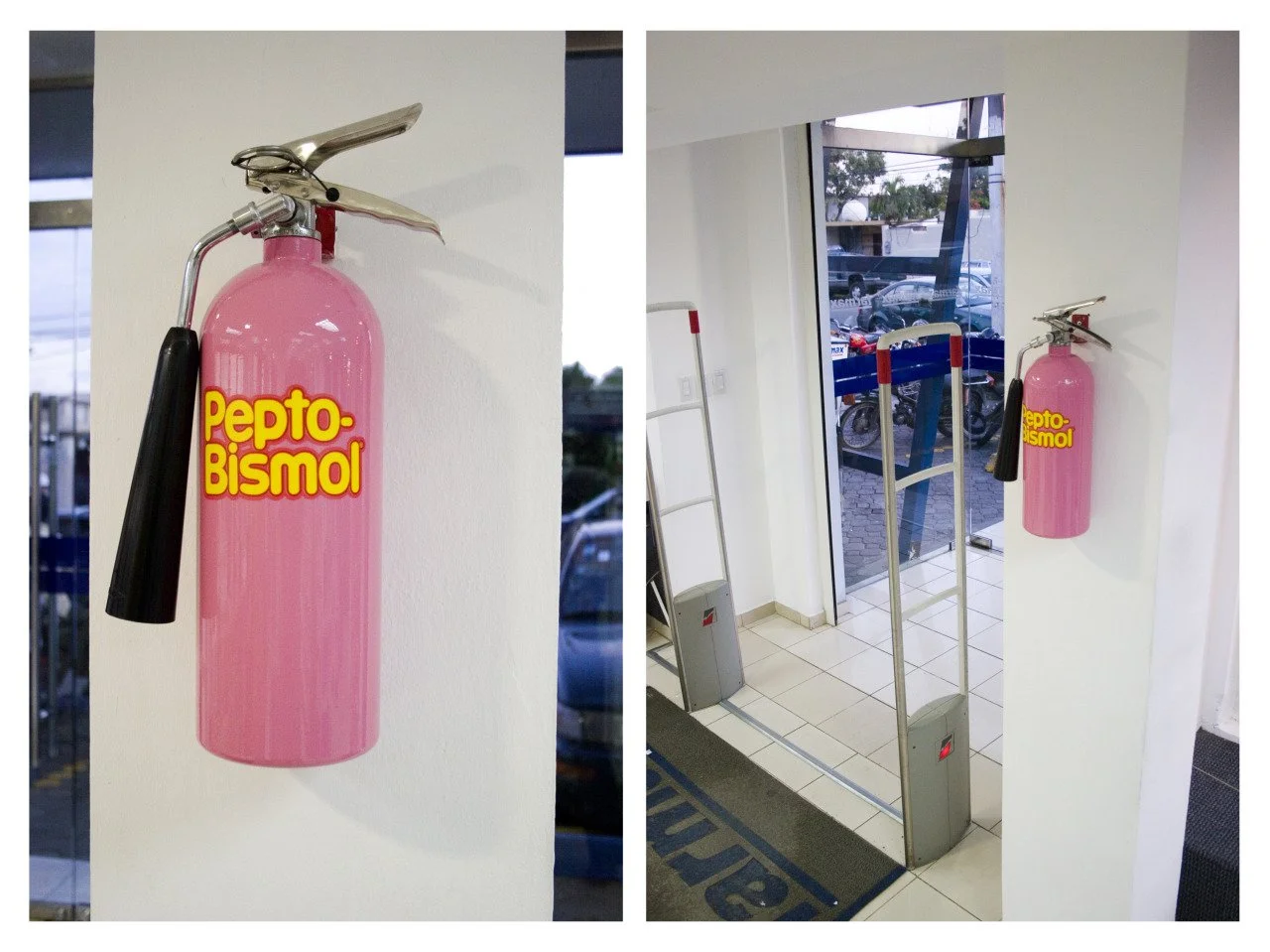 A pink fire extinguisher with a black nozzle mounted on a white wall near the entrance of a building. The view includes part of the doorway, a metal frame, and a glass door with a parking lot visible outside.