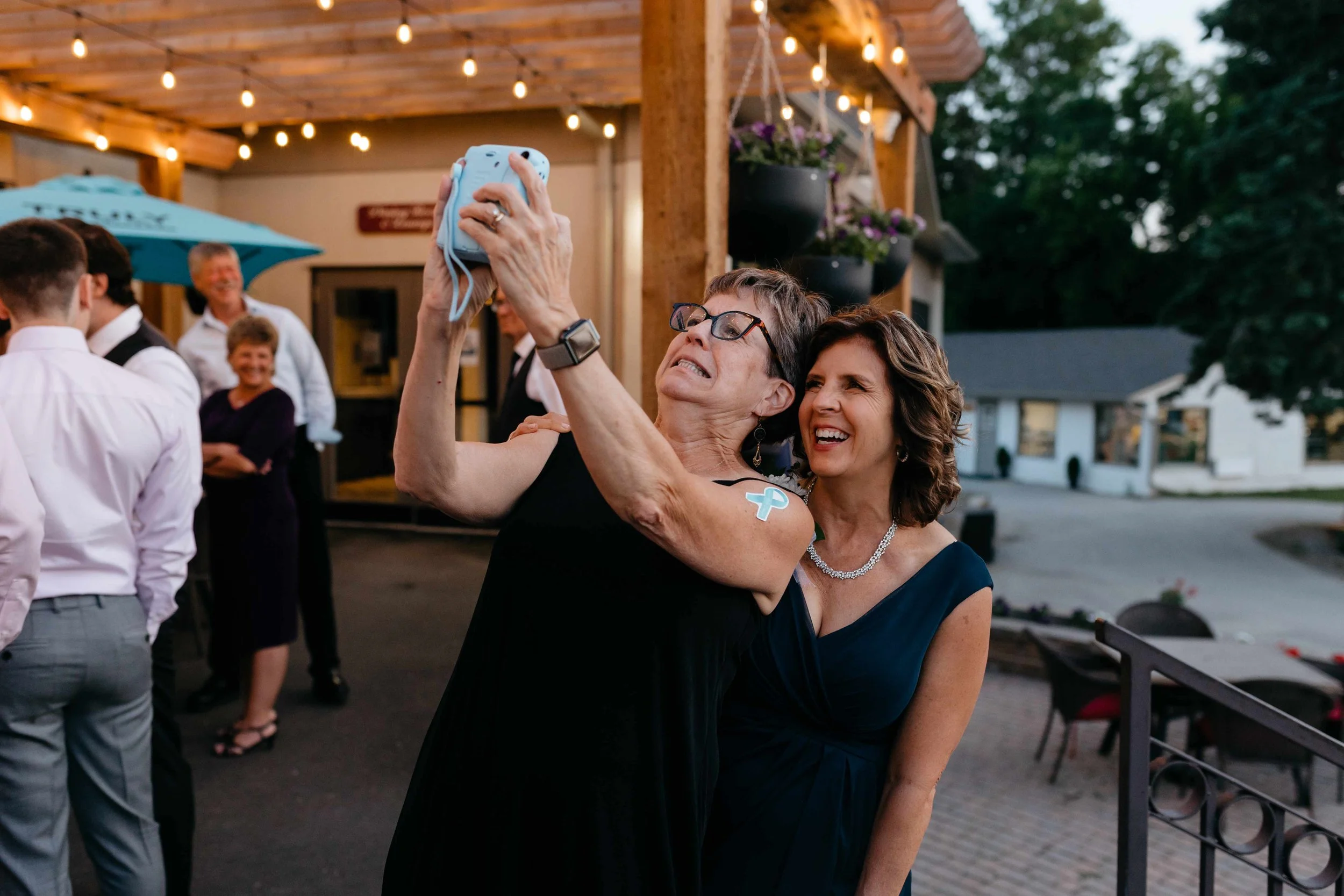 Two women taking a selfie at an outdoor event during the evening. One woman is wearing glasses, a black dress, and has a blue ribbon sticker on her arm; the other woman is smiling and wearing a dark dress with jewelry. There are string lights and oth
