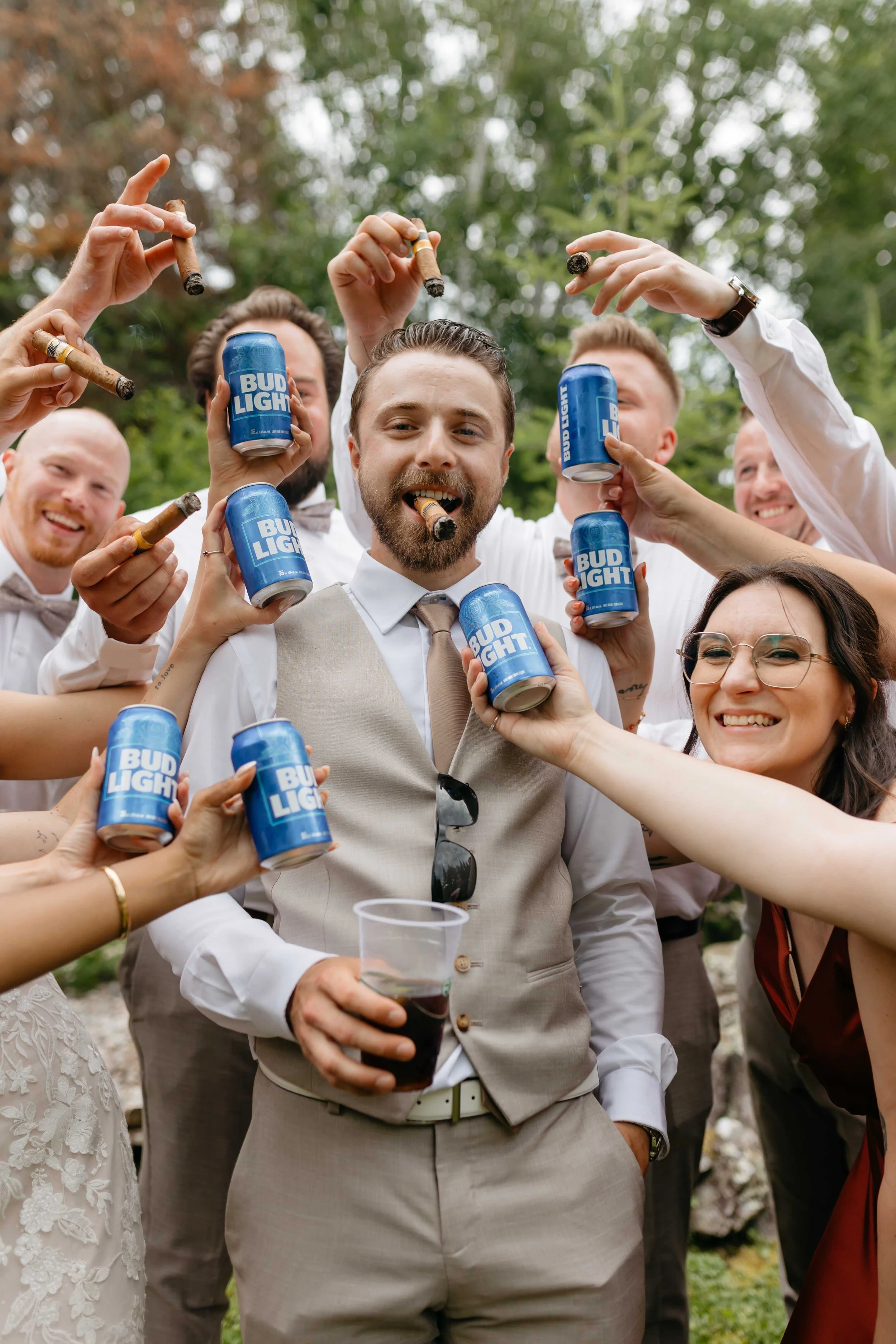 Group of friends at an outdoor celebration, holding Bud Light cans and cigars, dressed in semi-formal attire, with trees in the background.