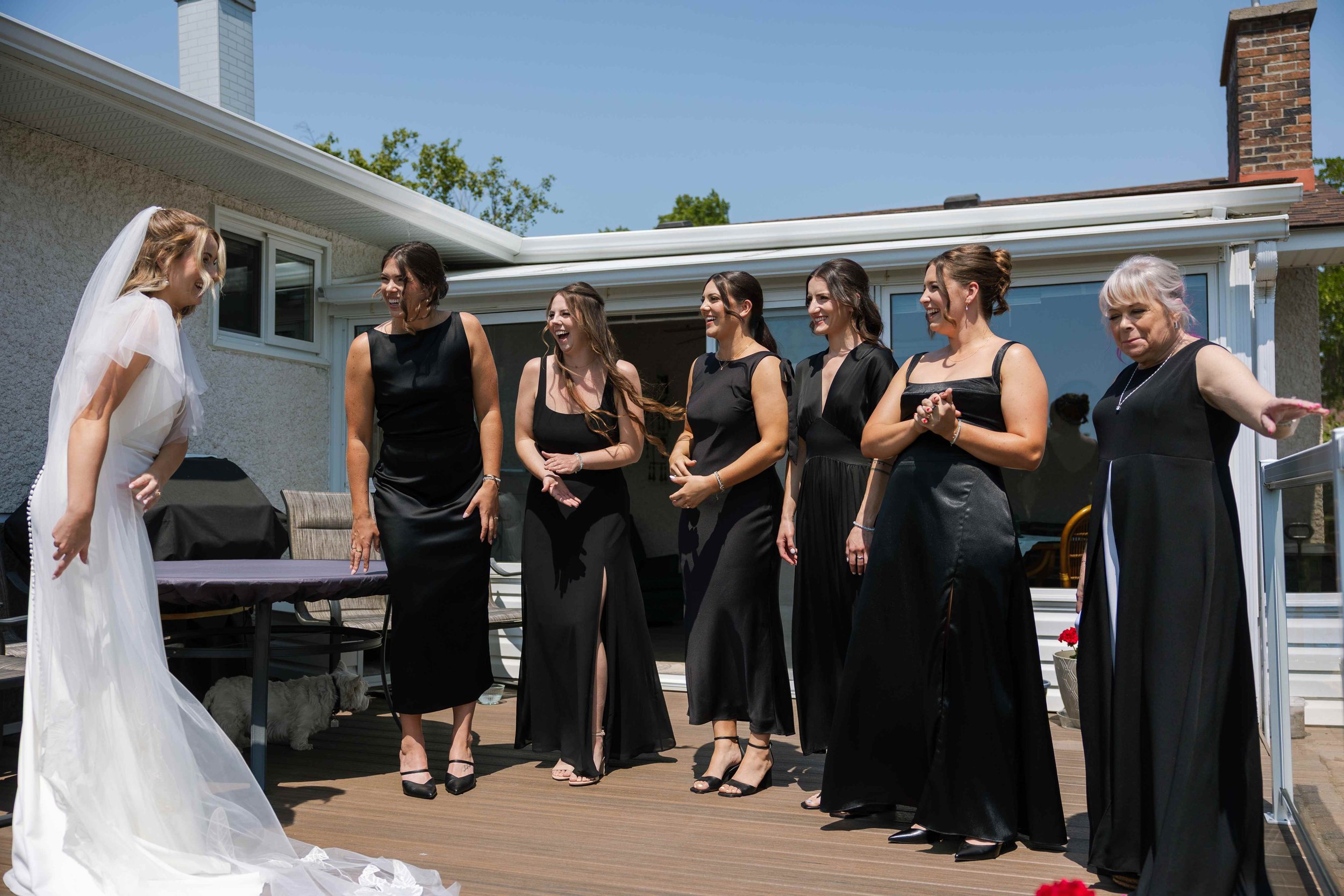 A bride in a white wedding dress and veil greeting a group of women dressed in black evening gowns at an outdoor wedding celebration on a sunny day.