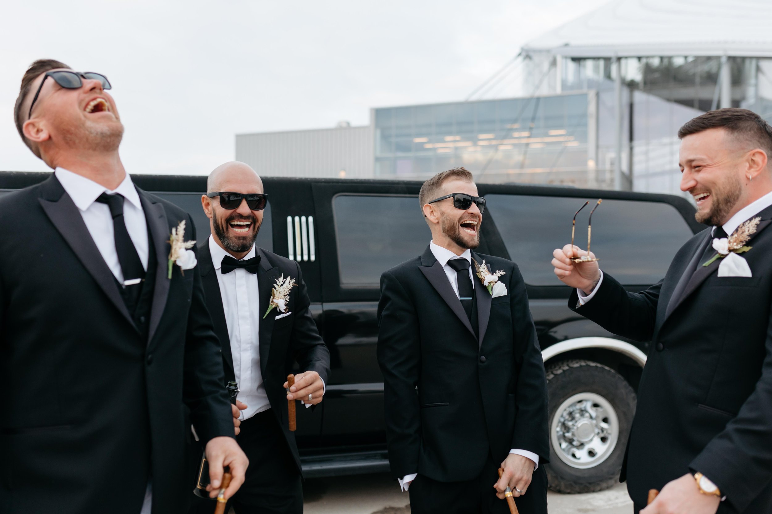 Group of men in black tuxedos and sunglasses standing outside near a luxury vehicle, smiling and laughing.