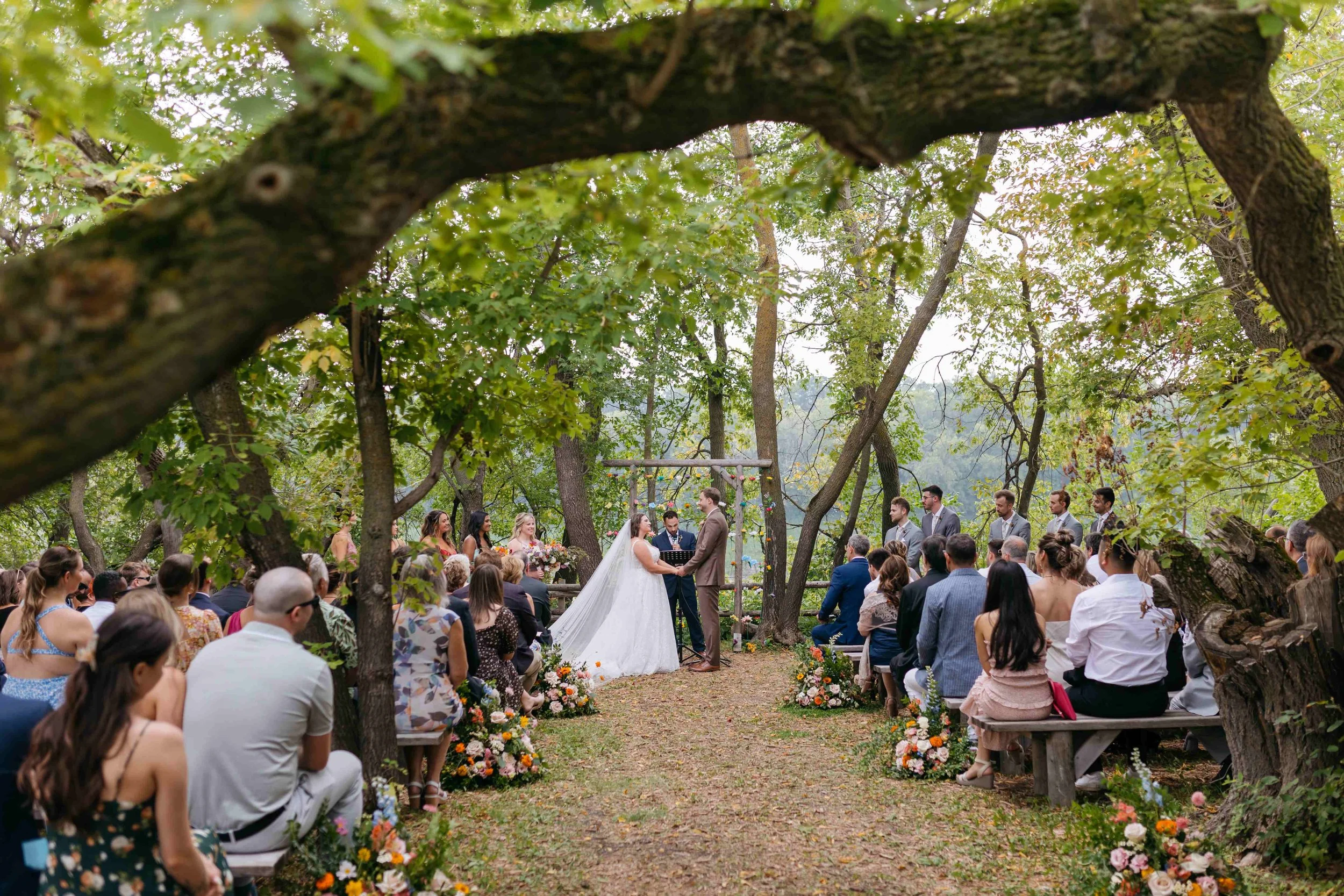 Outdoor wedding ceremony with a bride and groom exchanging vows under trees, surrounded by seated guests.