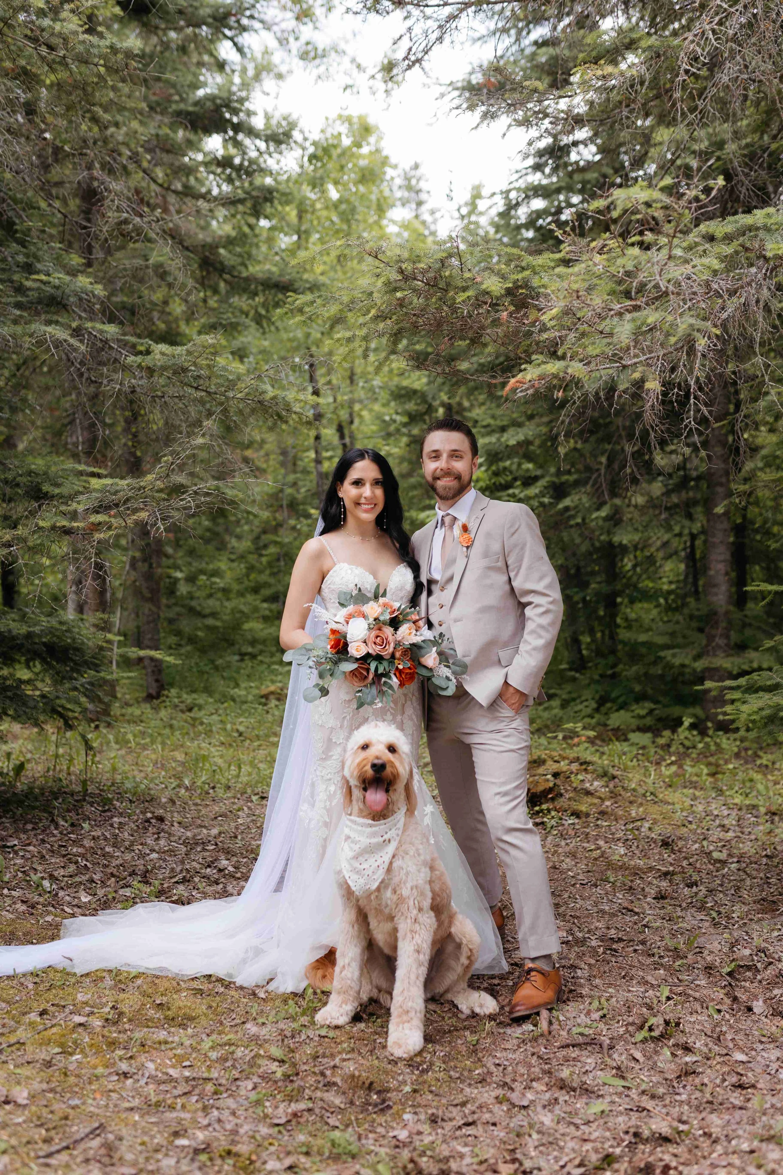 A bride and groom standing together outdoors in a forest, smiling, with a golden retriever sitting in front of them. The bride is wearing a white wedding gown and holding a bouquet, and the groom is dressed in a light gray suit. The dog wears a white