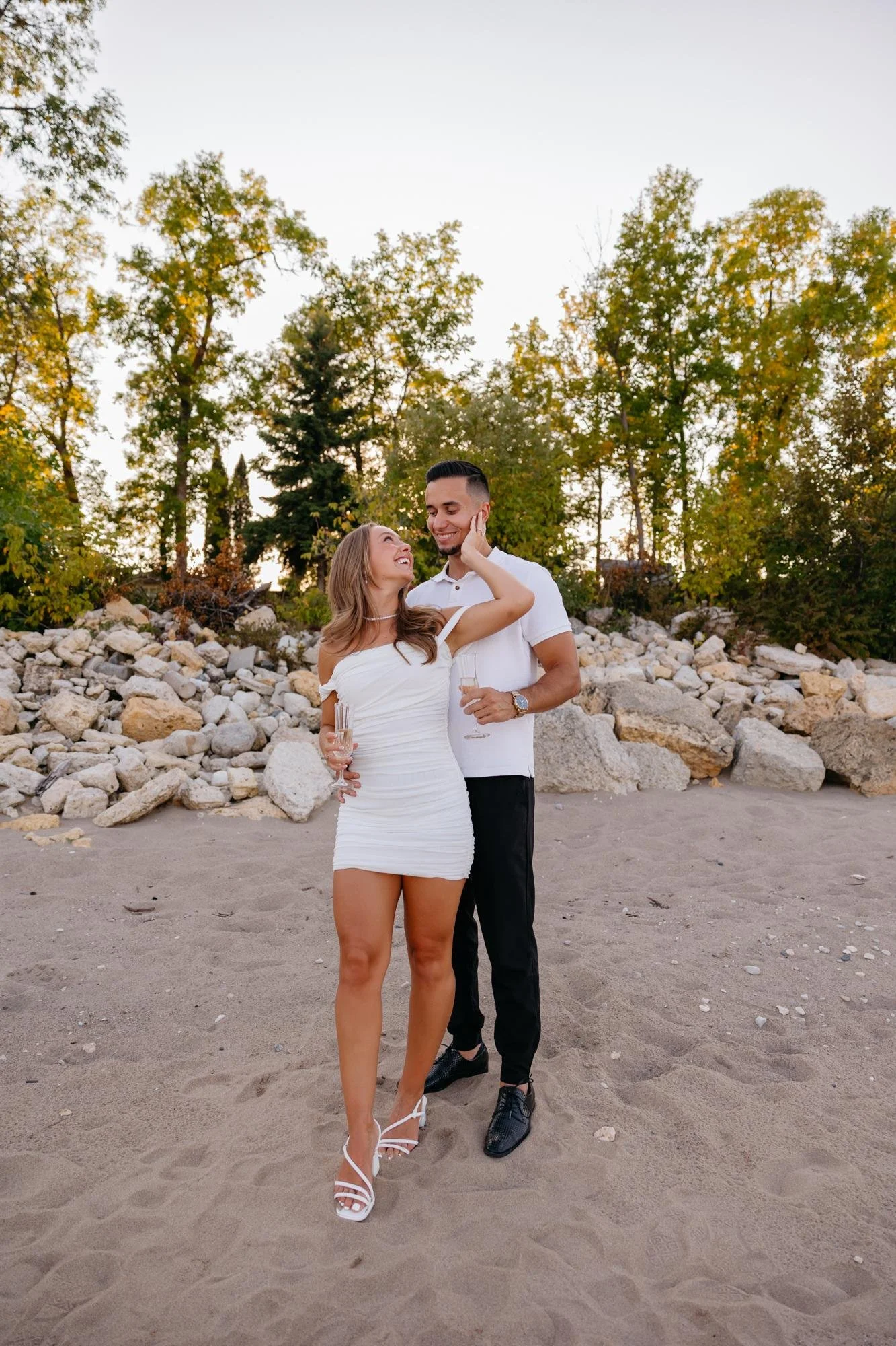 A couple standing on a sandy beach, smiling and looking at each other, with rocks and trees in the background, taken by Winnipeg couple photographer Sari Risbey.