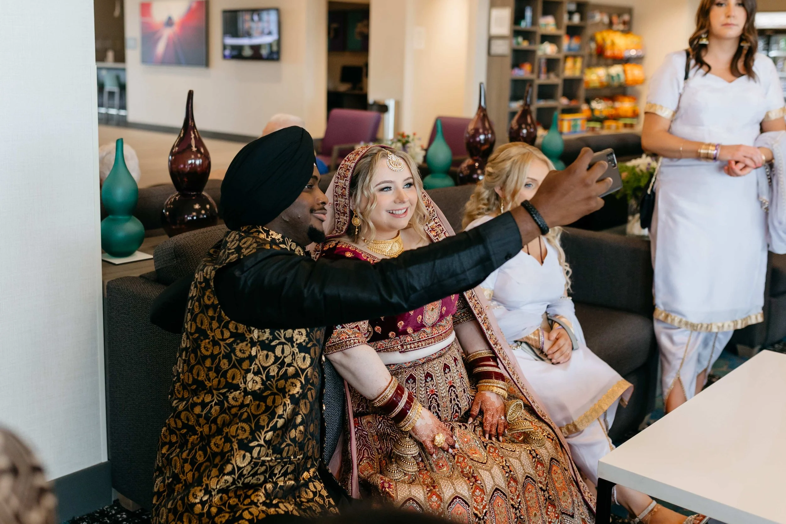 A group of diverse women dressed in traditional Indian attire taking a selfie together in a modern cafe or waiting area.