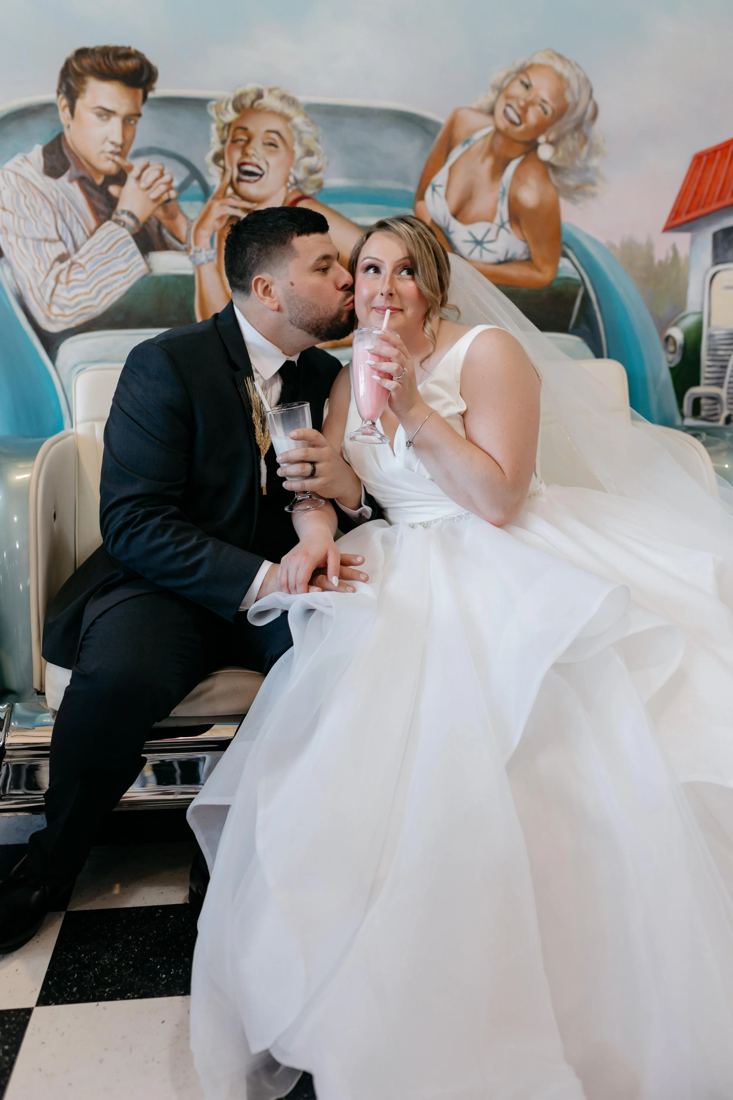 A newlywed couple sitting on a bench, with the groom kissing the bride's cheek as she drinks a pink milkshake. In the background, a mural of vintage-style women in a car is visible.