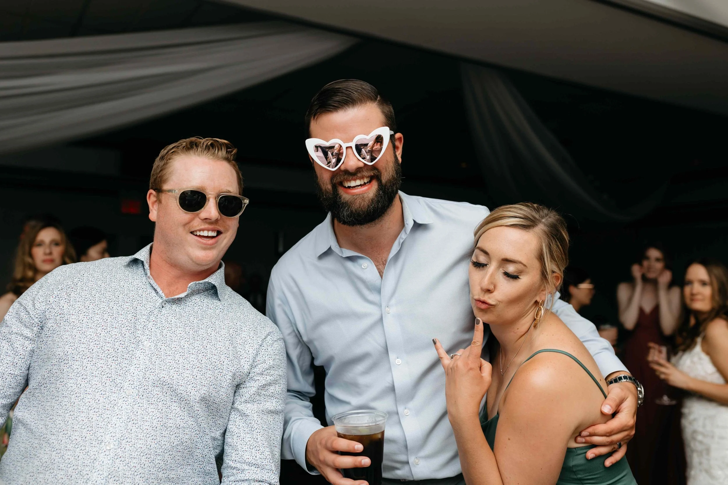 Three people smiling and posing for a photo at a social event. The man in the middle is wearing heart-shaped sunglasses and holding a drink, while the woman on the right is making a peace sign. The man on the left is wearing round sunglasses.