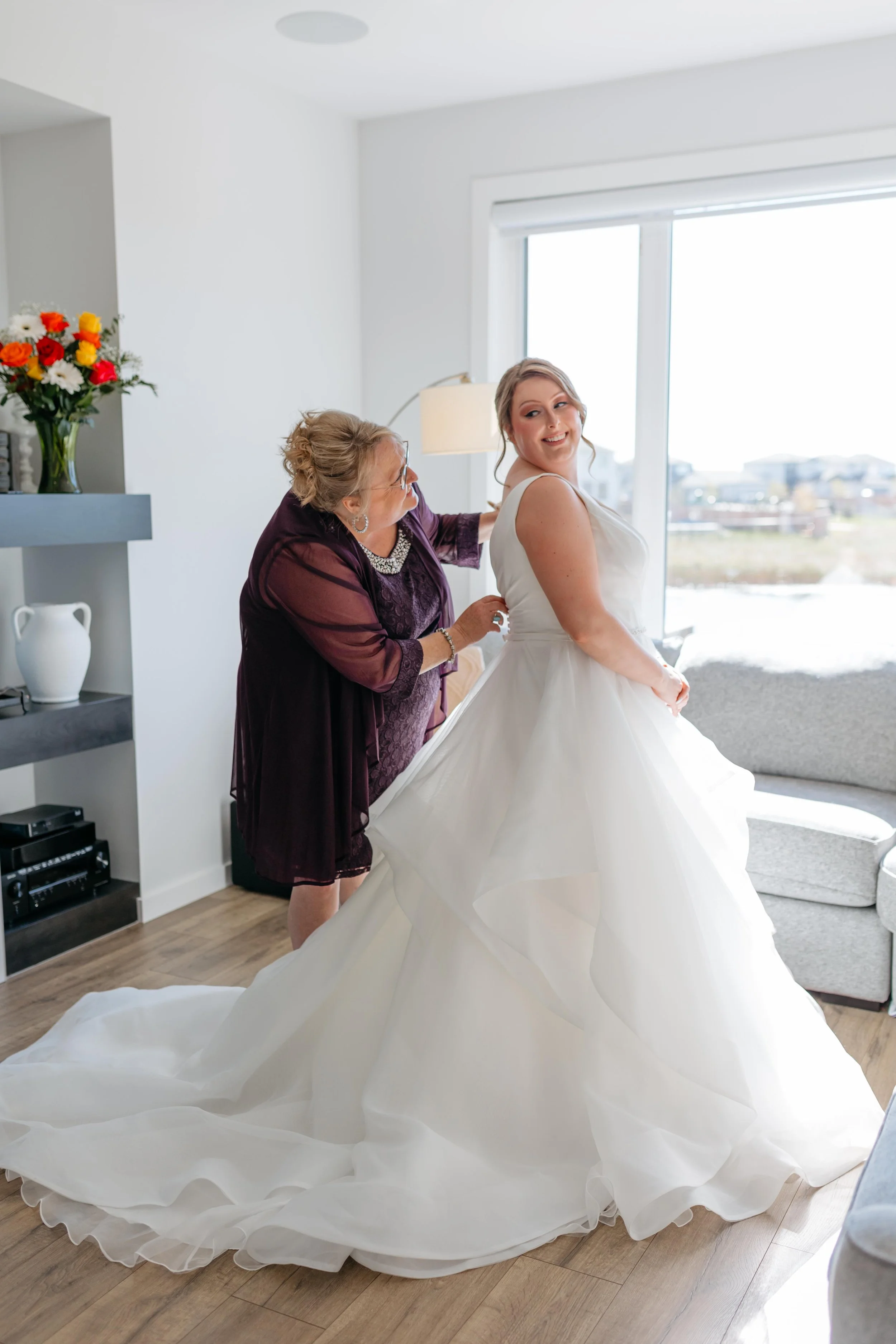 A bride in a white wedding gown being assisted with her dress by an older woman, in a bright room with large window, flowers, and furniture.