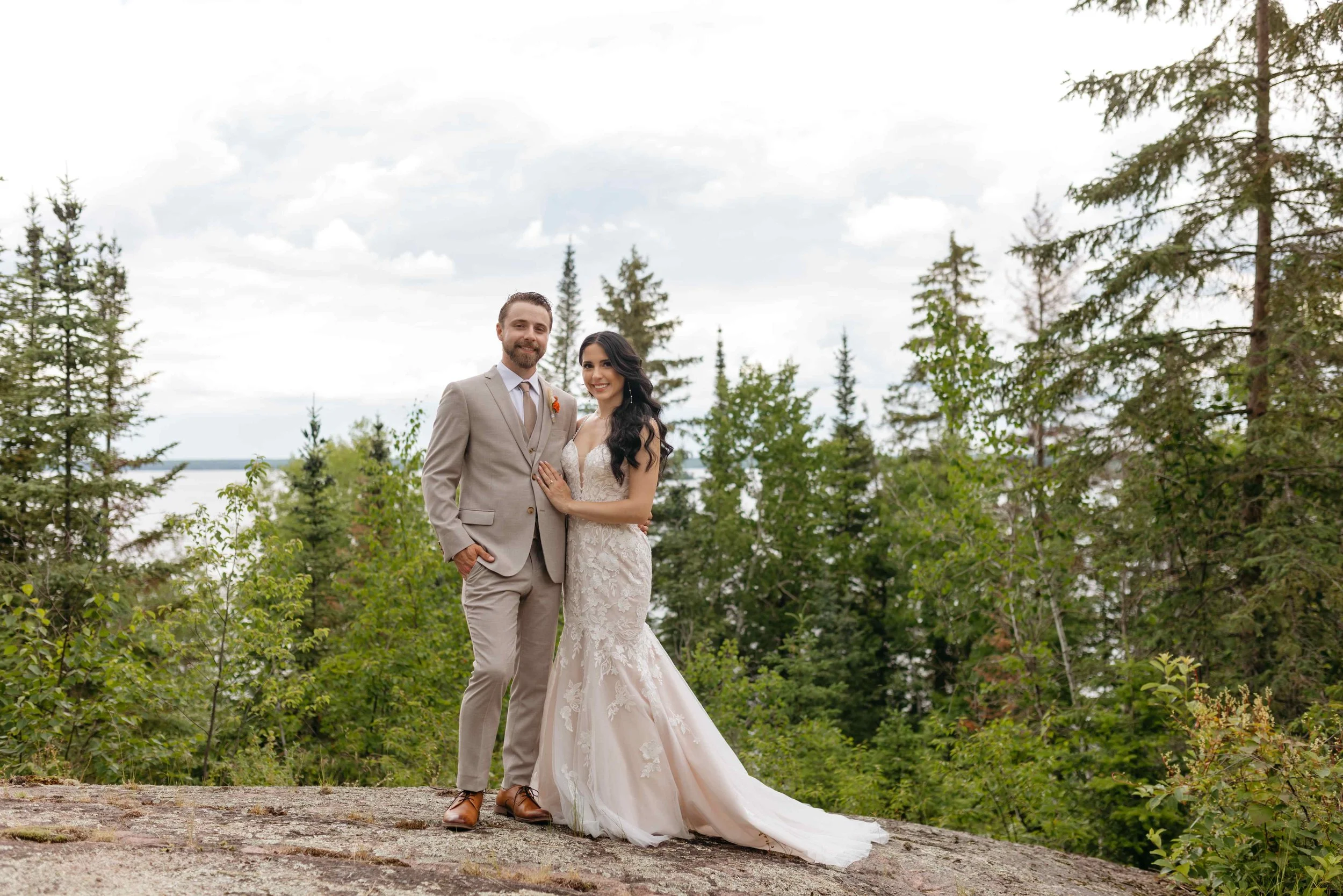 A bride and groom standing outdoors on a rocky surface with trees and a cloudy sky in the background. The groom is wearing a beige suit and the bride is in a white lace wedding dress. They are smiling and close to each other.