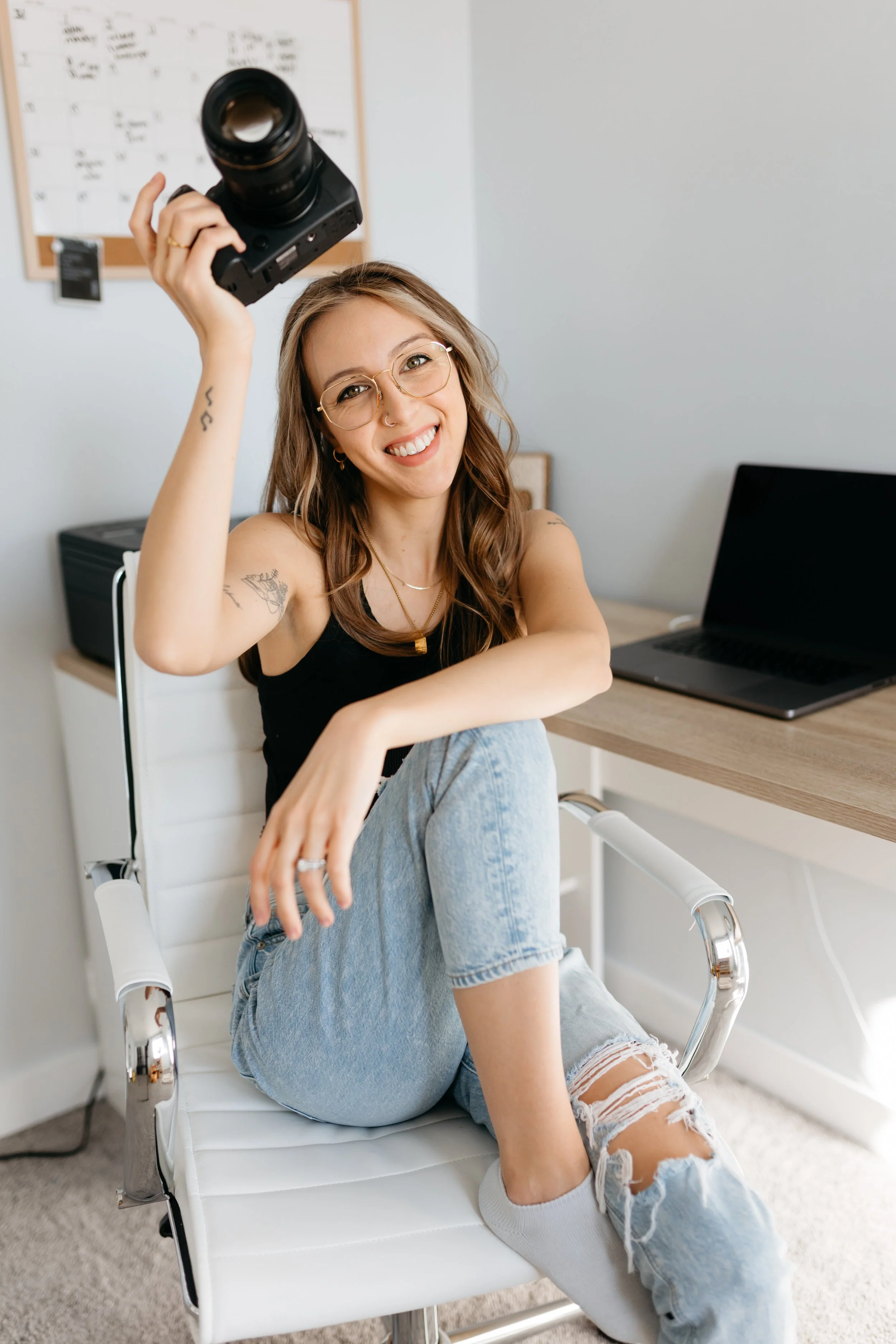 Sari Riseby, a Winnipeg family and newborn photographer, is holding a camera above her head while sitting on a modern white chair.