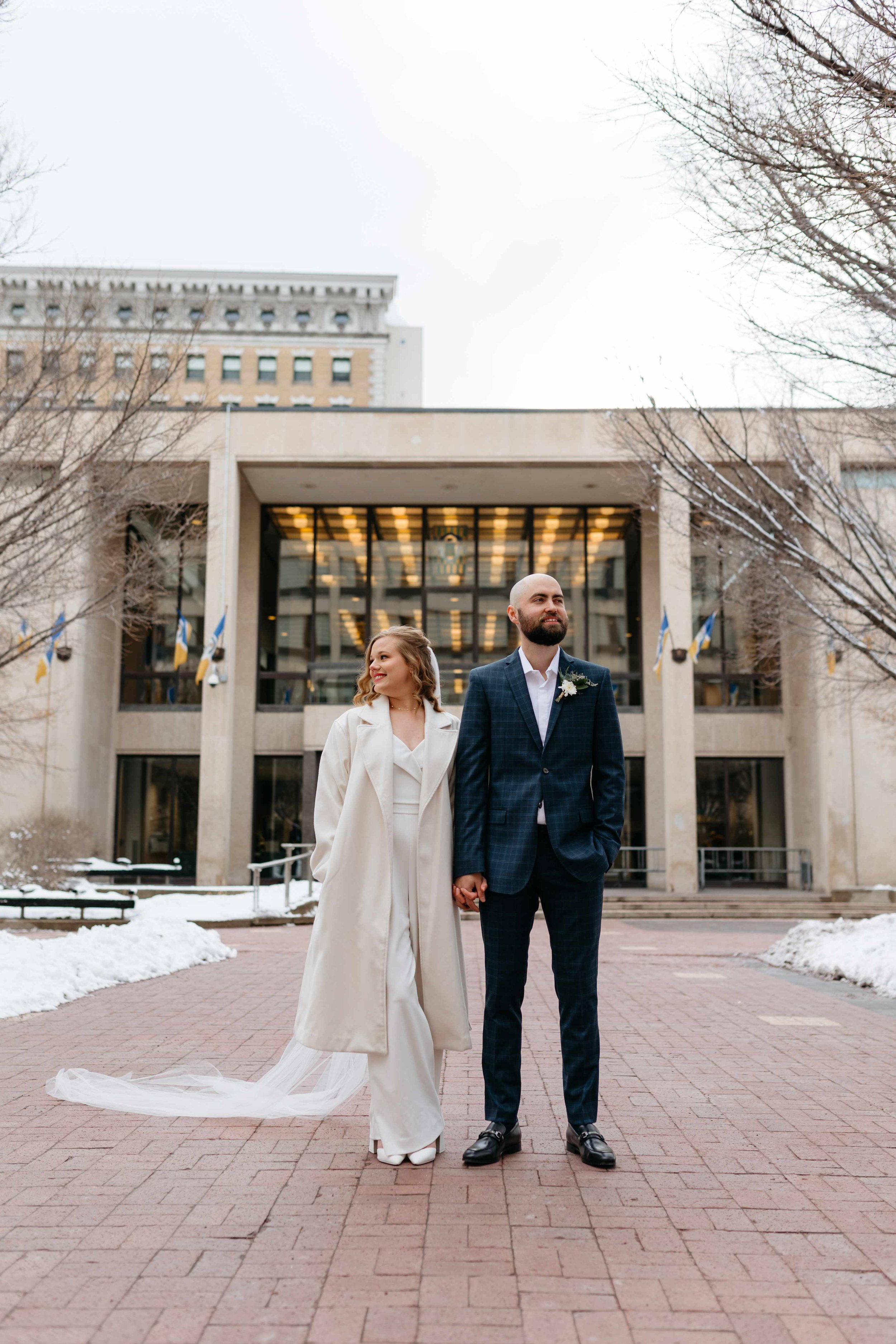 A bride and groom standing outside in a winter setting, holding hands, in front of a government building with flags, trees with no leaves and snow on the ground.