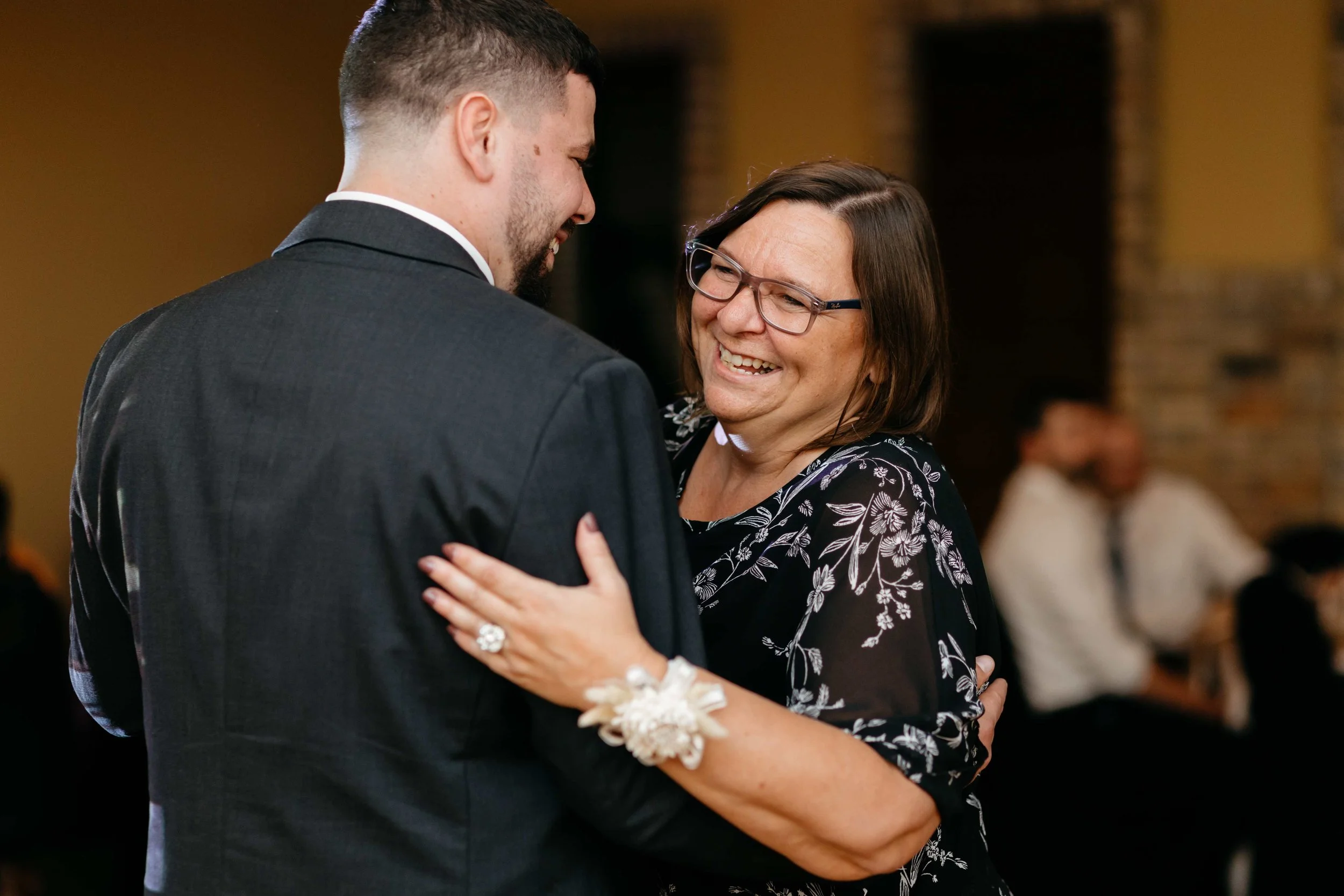A man and a woman are dancing closely, smiling and embracing each other at a social event, with a blurred background and warm lighting.