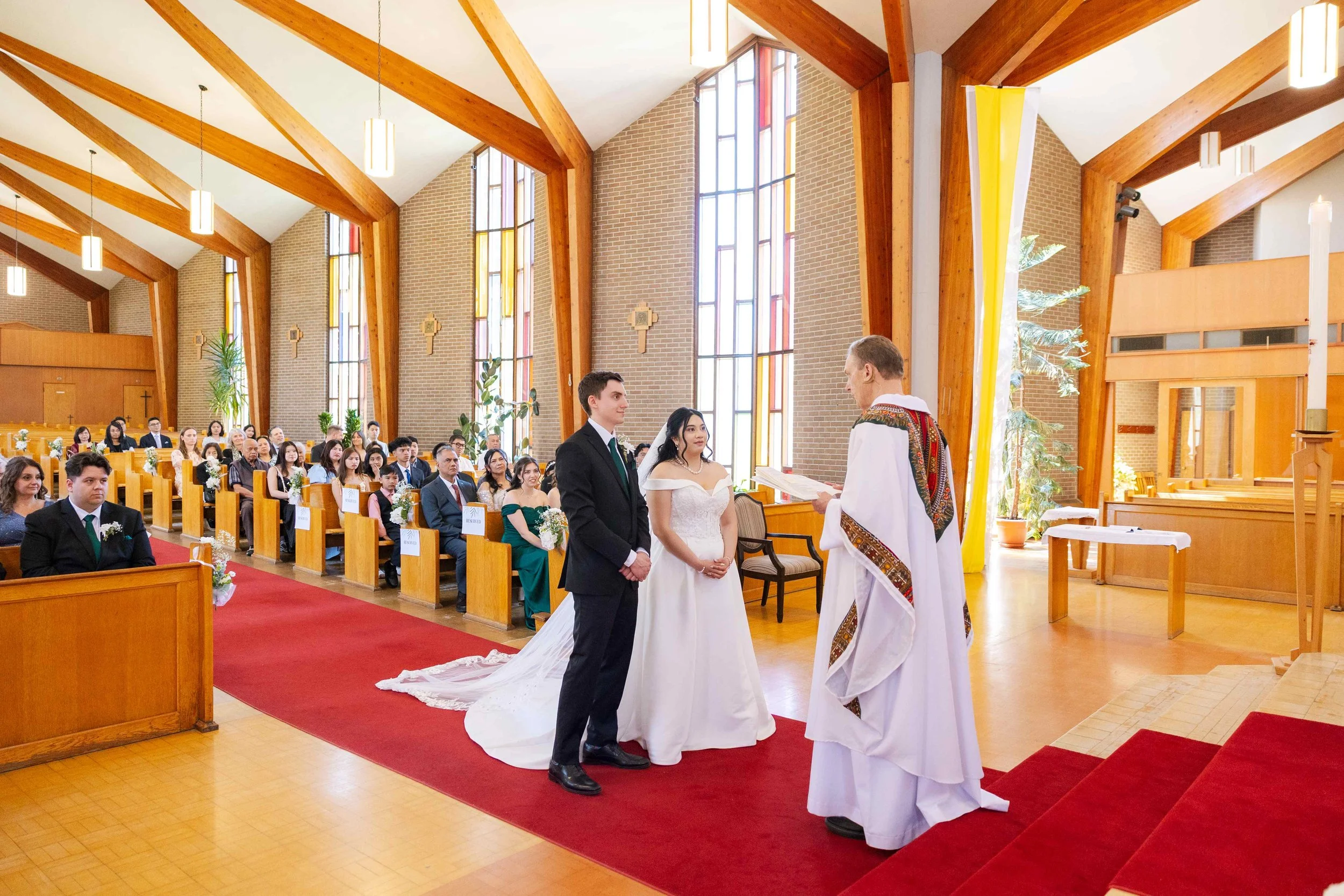 A wedding ceremony inside a church with a bride and groom standing before a priest, with guests seated in pews behind them.