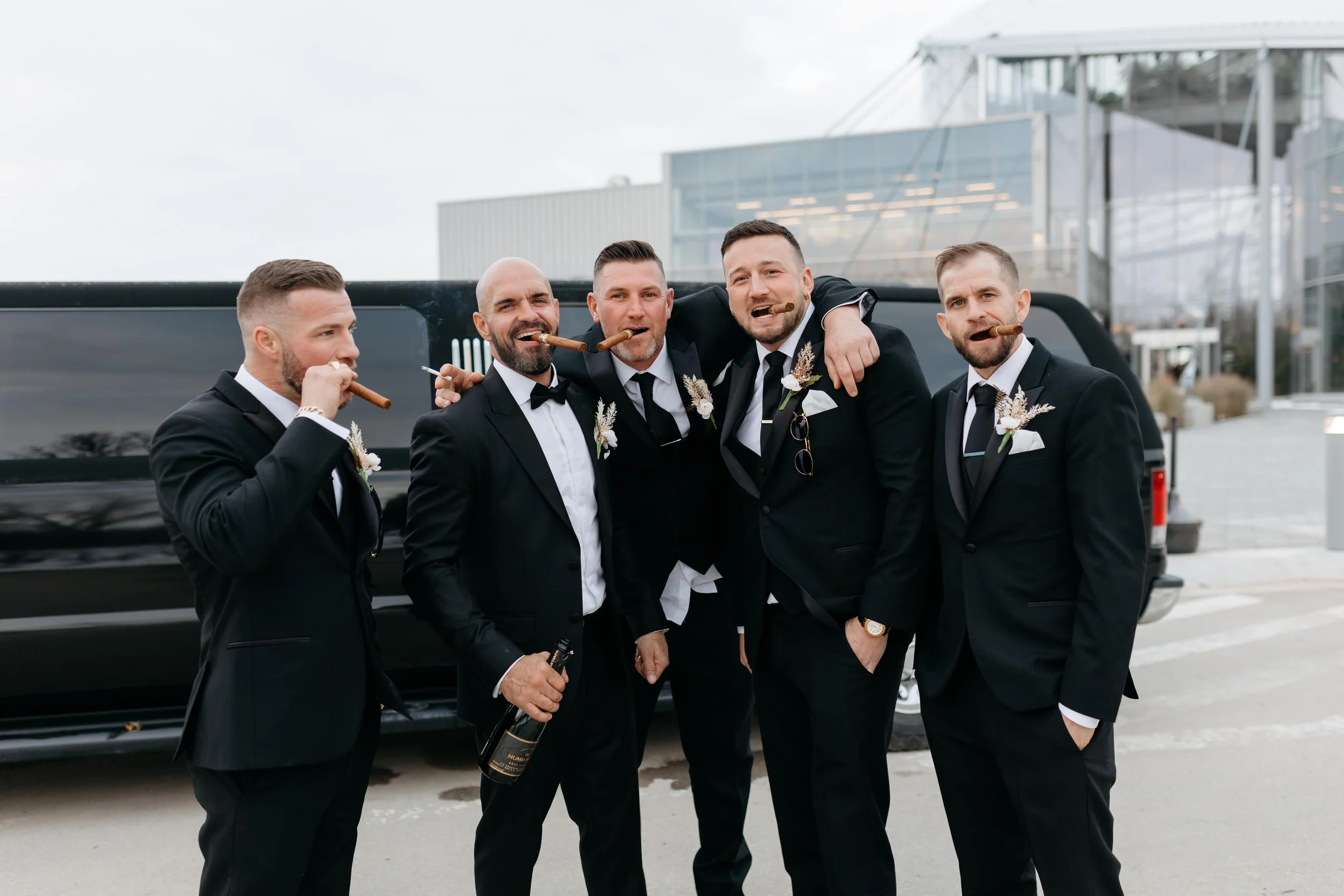 Five men dressed in black tuxedos with white shirts and black bow ties, celebrating outdoors in front of a black limousine, smoking cigars, and holding a bottle of champagne. They appear happy and are smiling, with one having his arm around another.