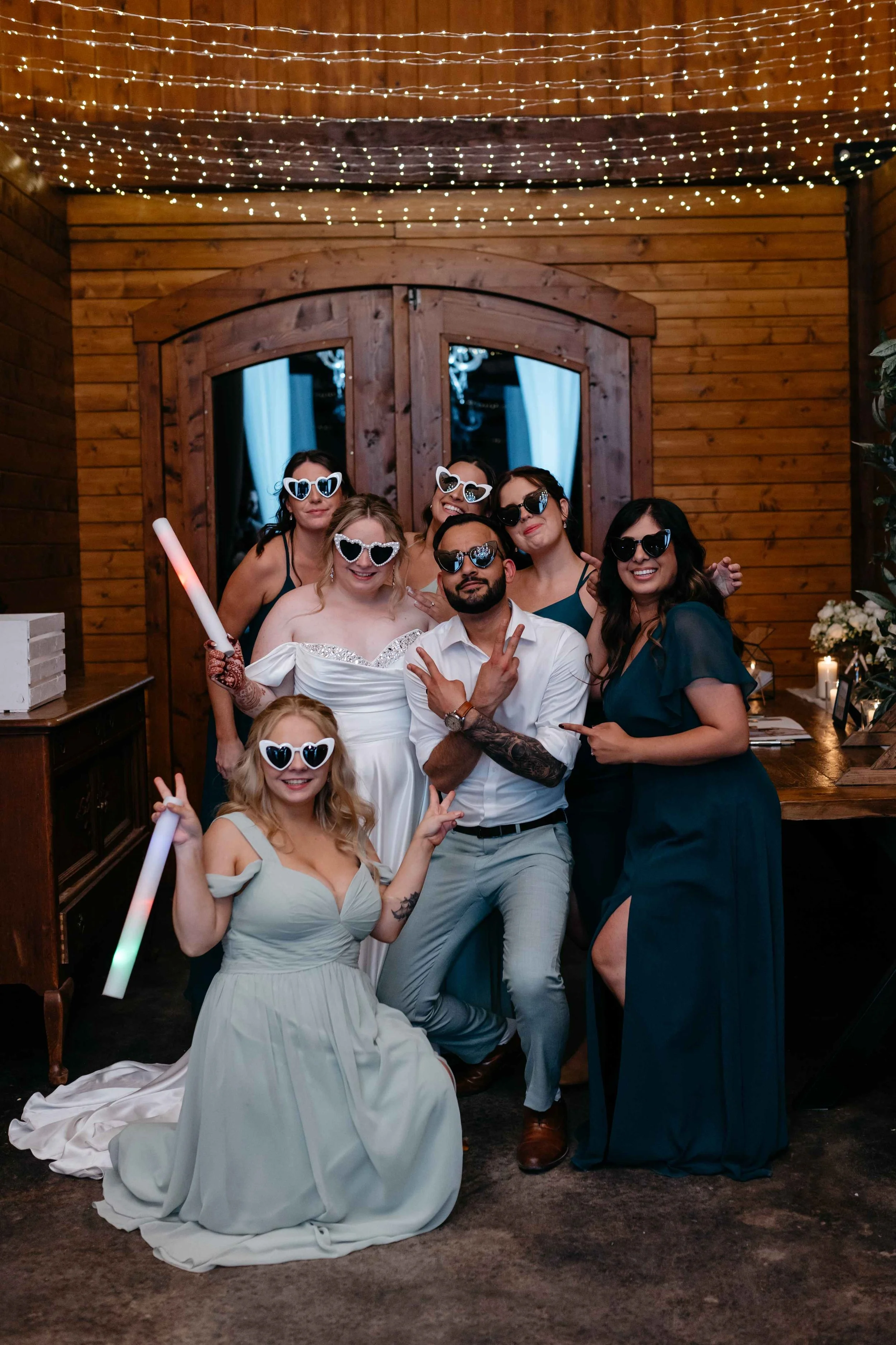 Group of people celebrating at a party, wearing heart-shaped sunglasses and holding glow sticks, with a wooden wall and string lights in the background.