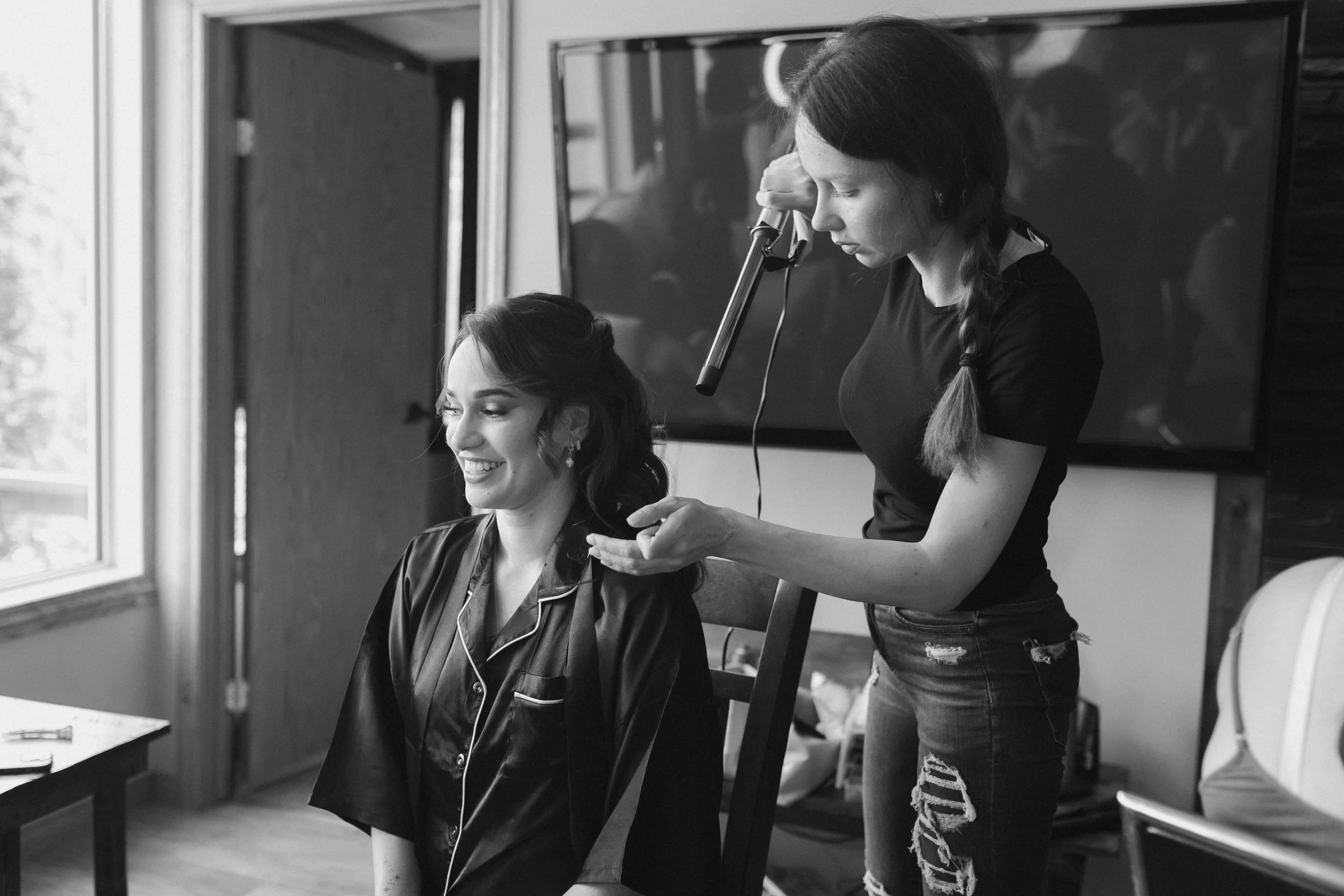 A woman is sitting in a chair getting her hair styled by a hairdresser in a dressing room, with a window and a television screen in the background.