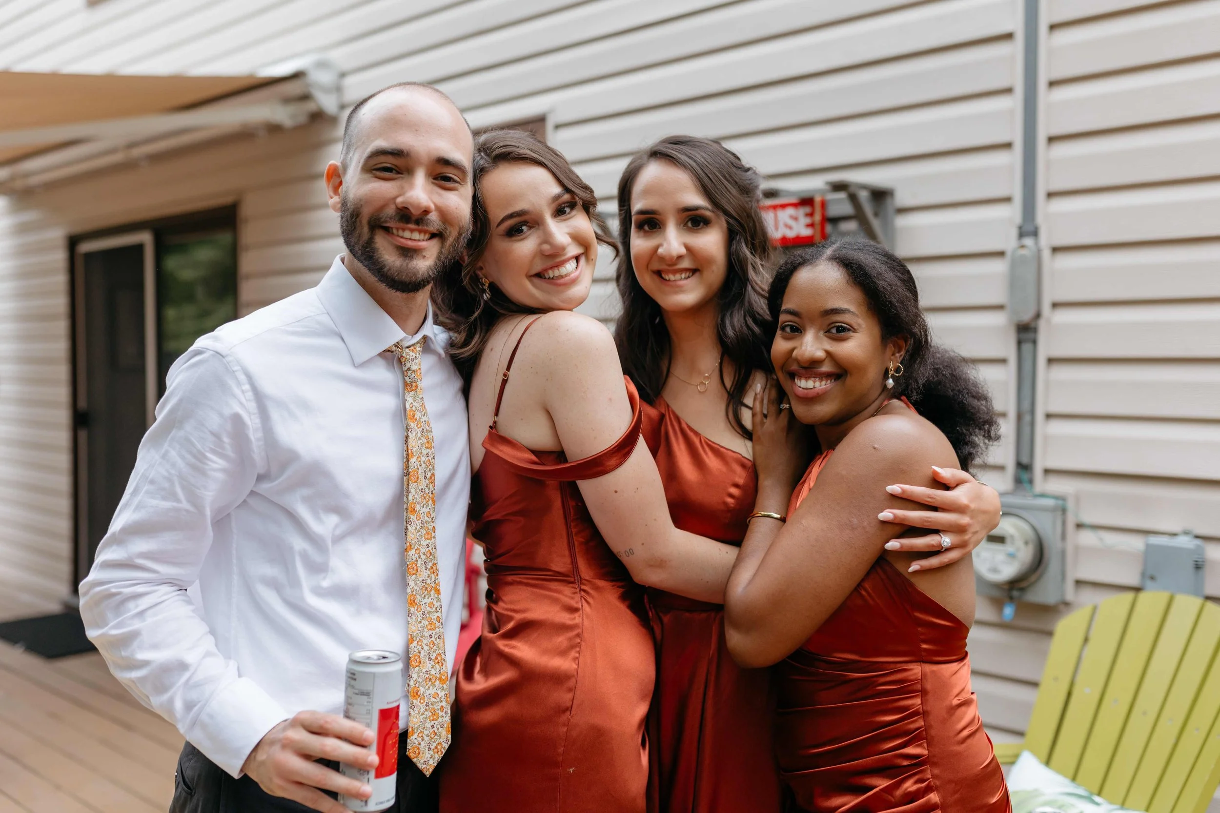 Four friends, three women and one man, smiling and hugging at an outdoor gathering, with a beige siding building in the background.