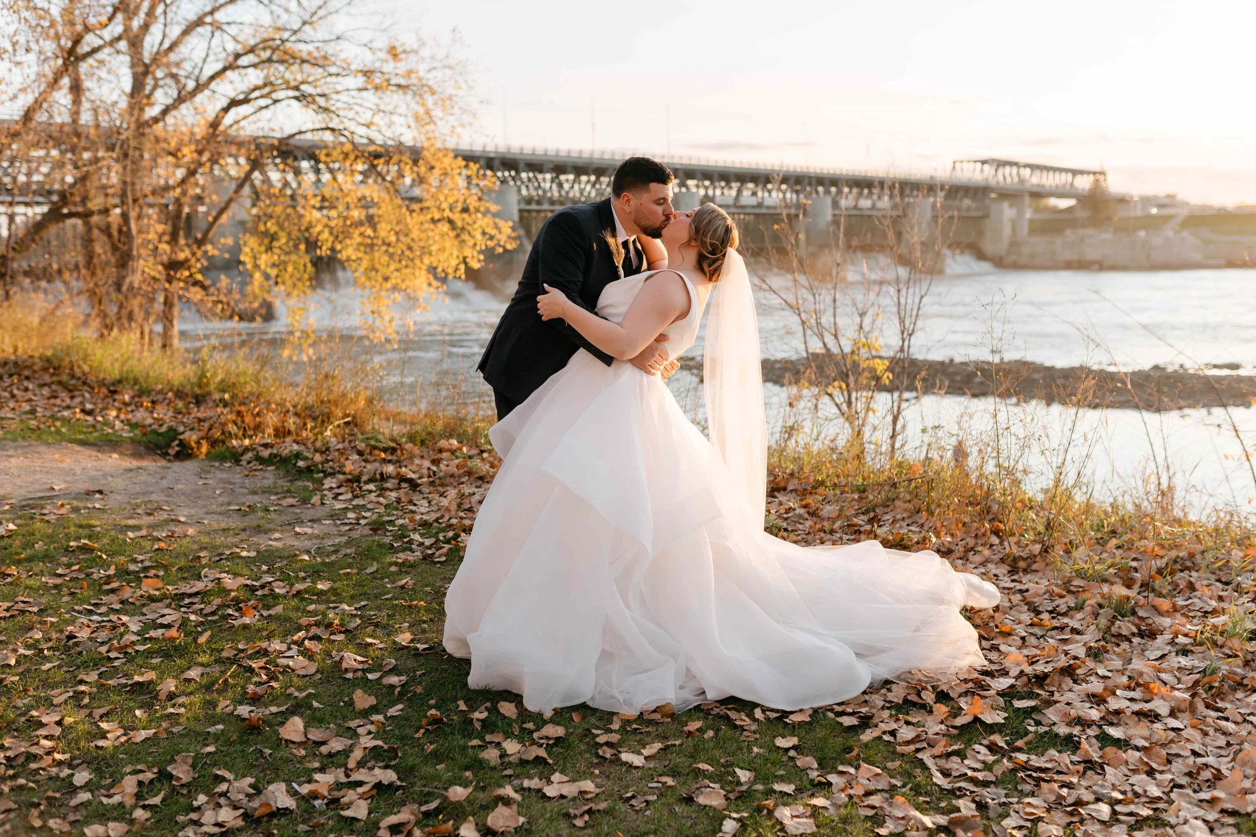 A bride and groom sharing a kiss outdoors near a river, with autumn leaves on the ground and a bridge in the background during sunset.