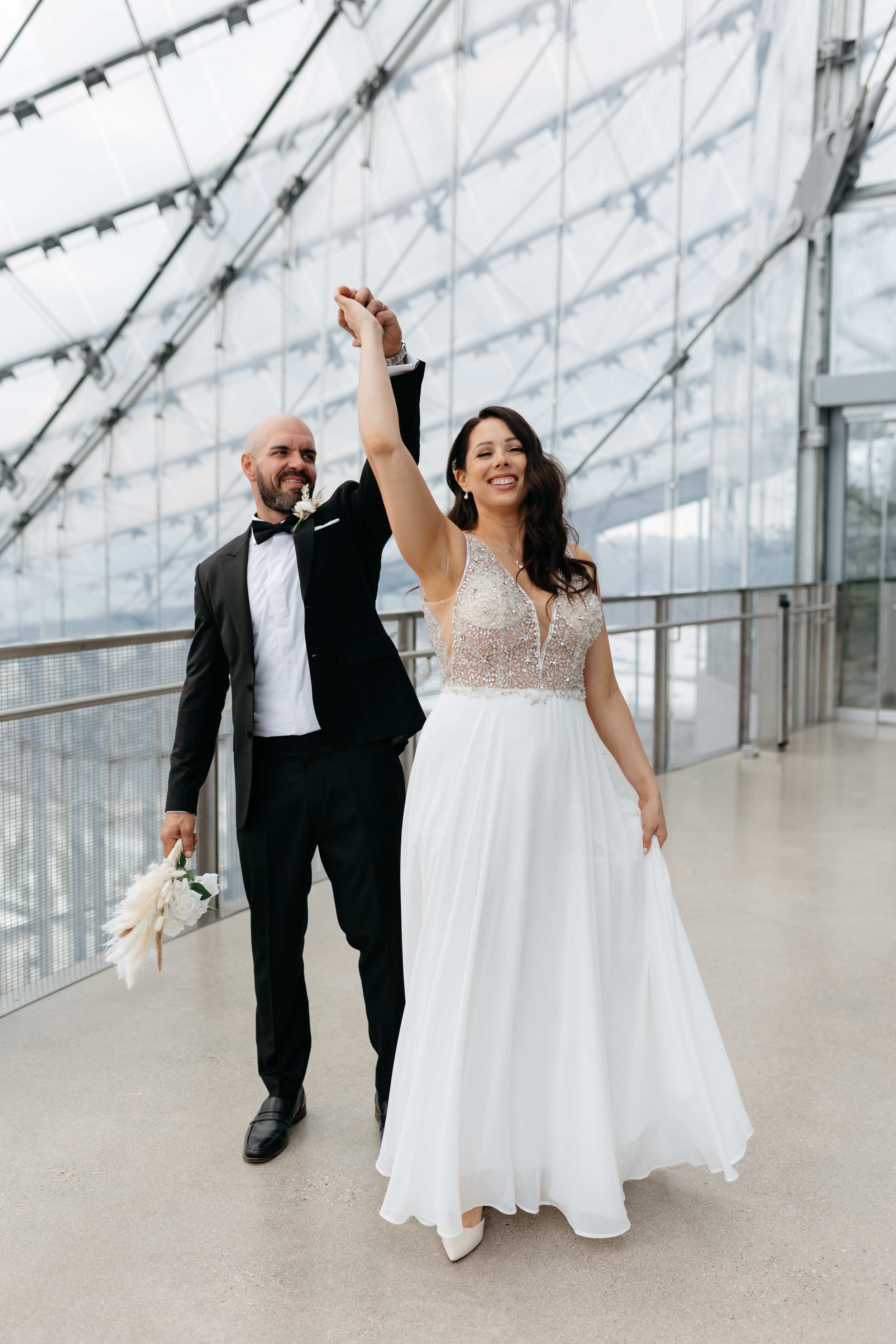 A newlywed couple holding hands and smiling as they walk through an indoor space with large glass windows and metal framework in Winnipeg