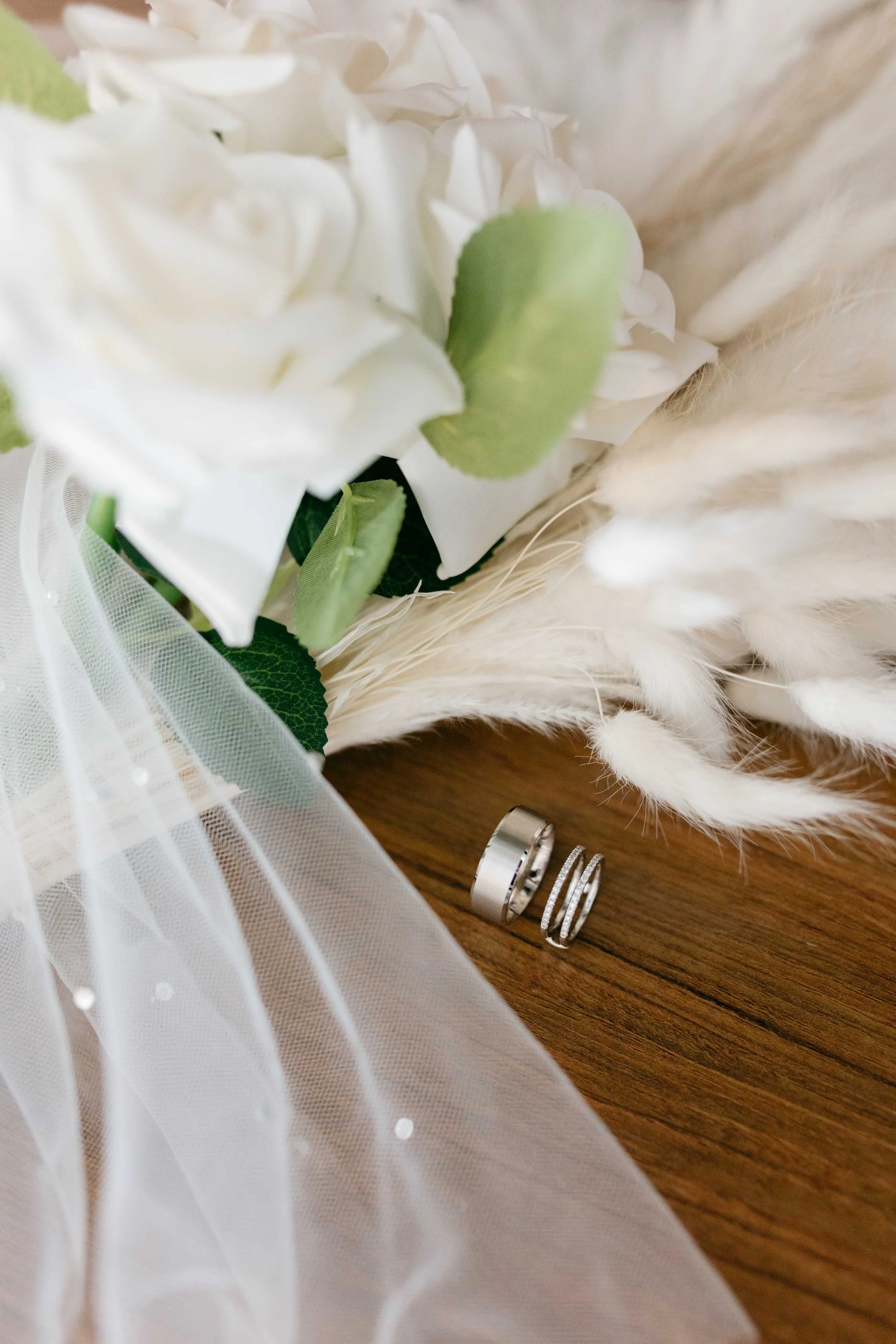 Close-up of wedding rings on a wooden surface, decorated with white lace, green leaves, white flowers, and soft feathers.